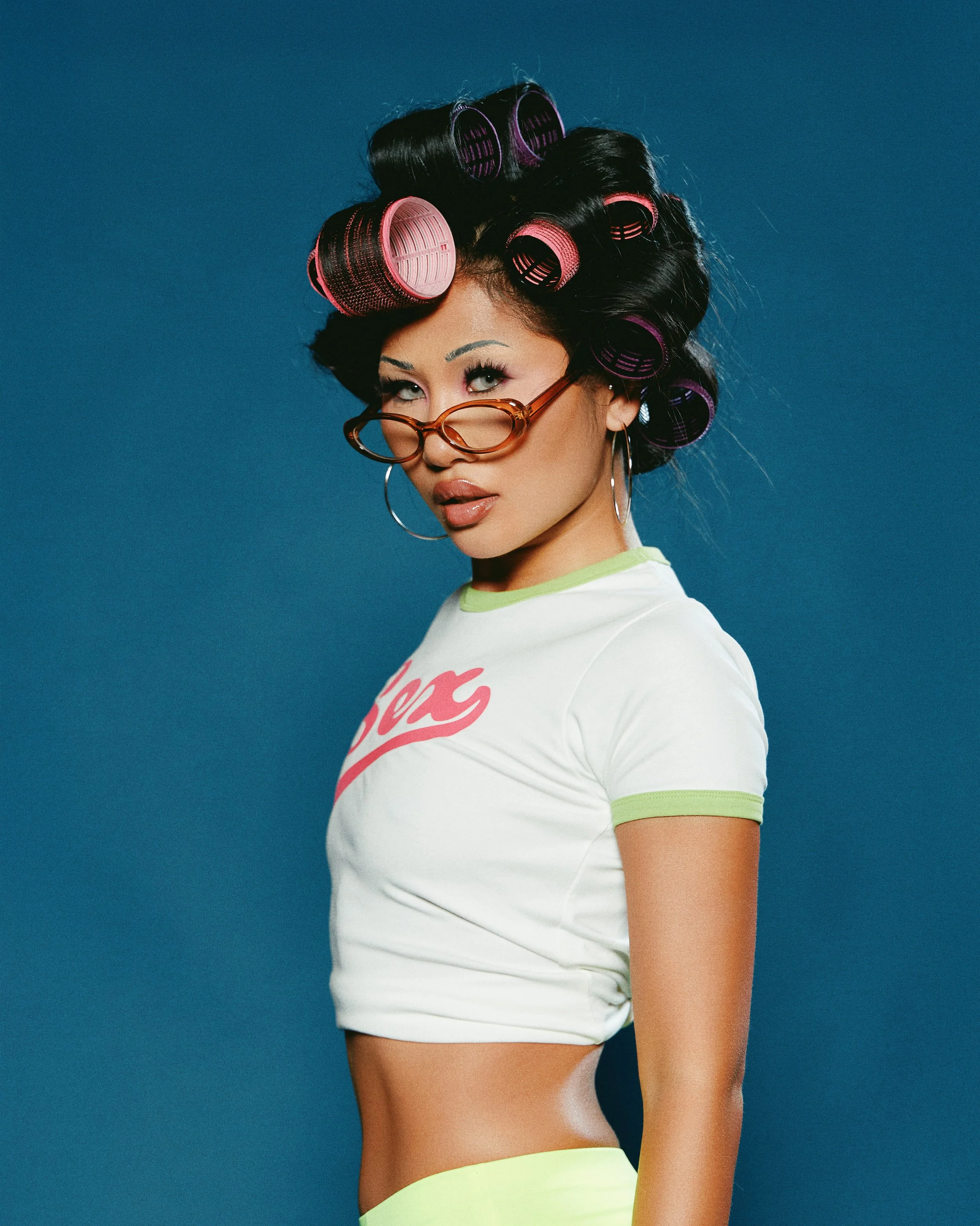 A woman with hair rollers in her hair, wearing glasses, hoop earrings, and a white t-shirt with pink writing, posing against a blue background.