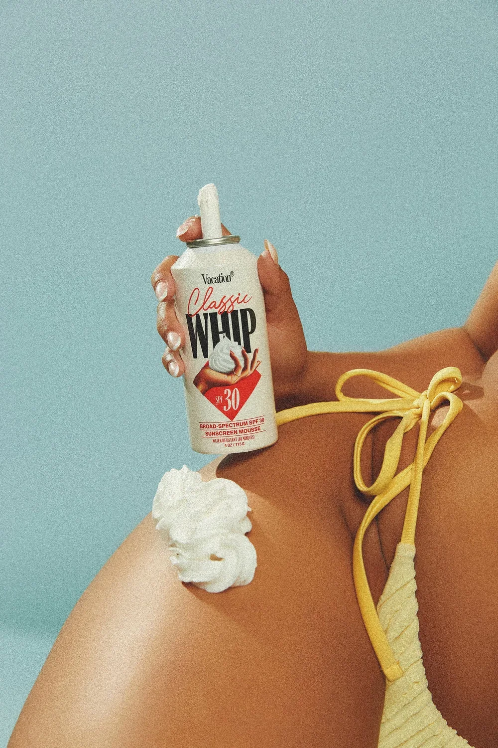 A person holding a bottle of sunscreen mousse labeled 'Vacation Classic Whip SPF 30' with a dollop of the white foam on their thigh, wearing a yellow swimsuit tied with bow on the side, against a blue background.