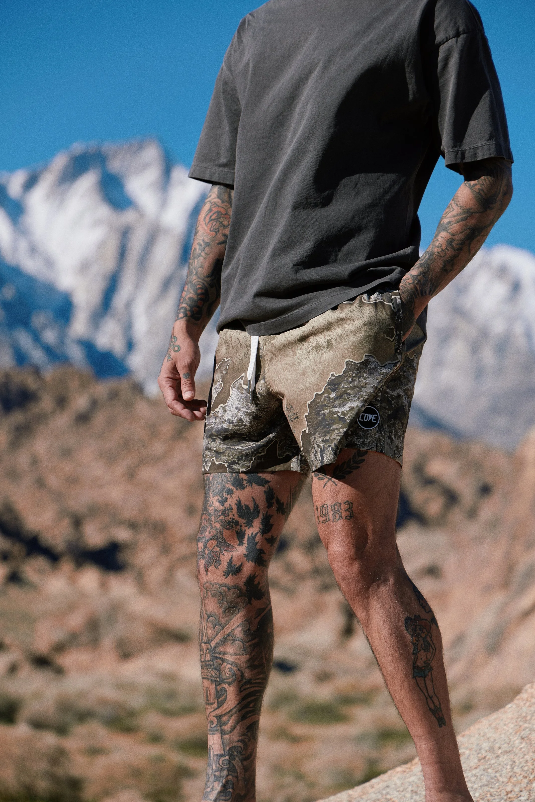 Activewear photography in Los Angeles: camo shorts with snowy mountains behind, low angle, by Moises Martinez