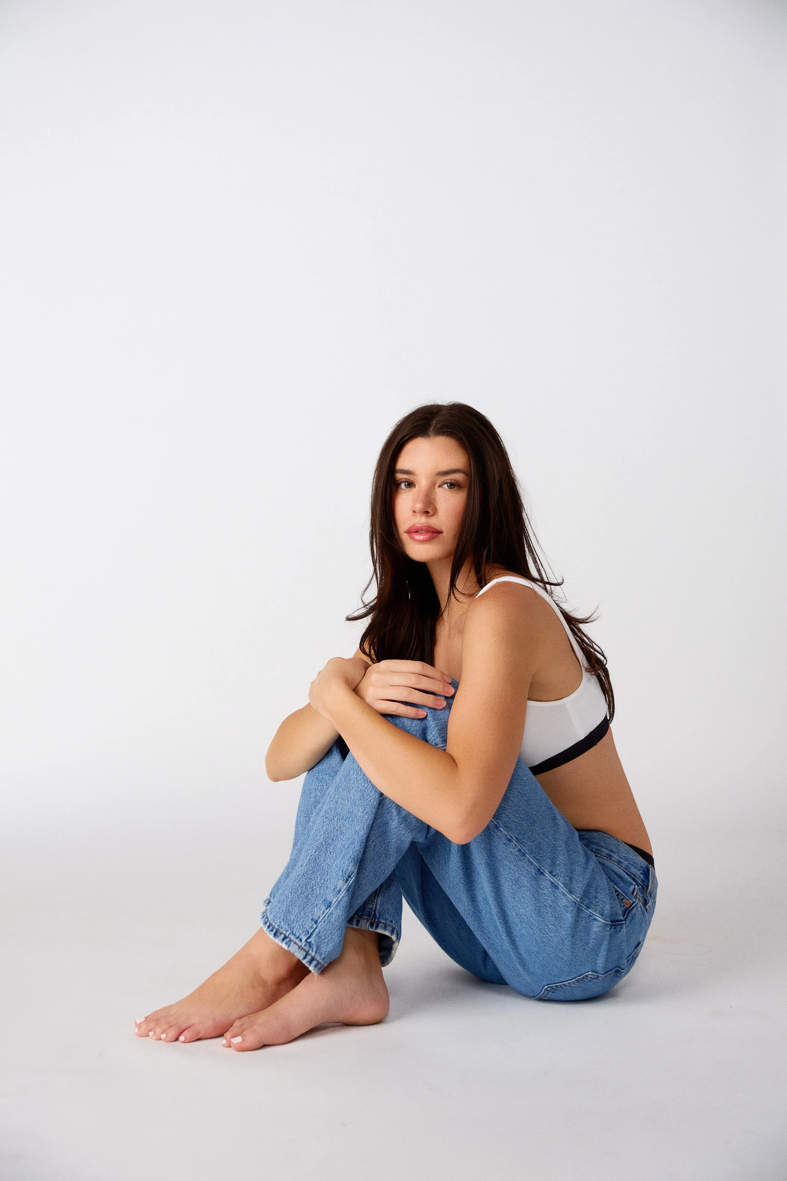 A young woman with long brown hair sitting on the floor, hugging her knees. She is wearing a white and black sports bra and blue jeans, with a neutral expression against a plain white background.