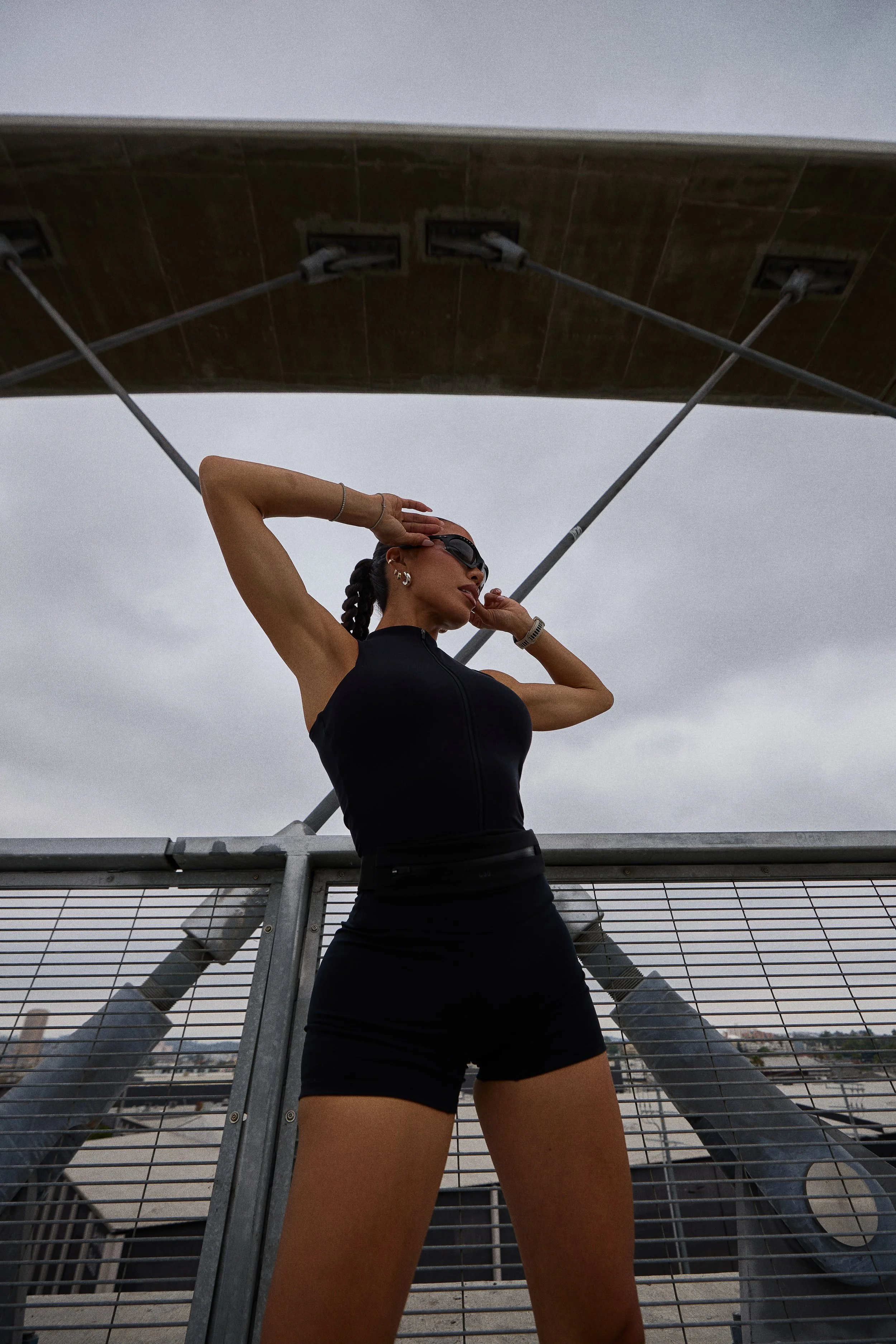 A woman in black athletic wear and sunglasses stands on a rooftop with a metal railing, striking a pose with one arm behind her head and the other touching her ear, against a cloudy sky.