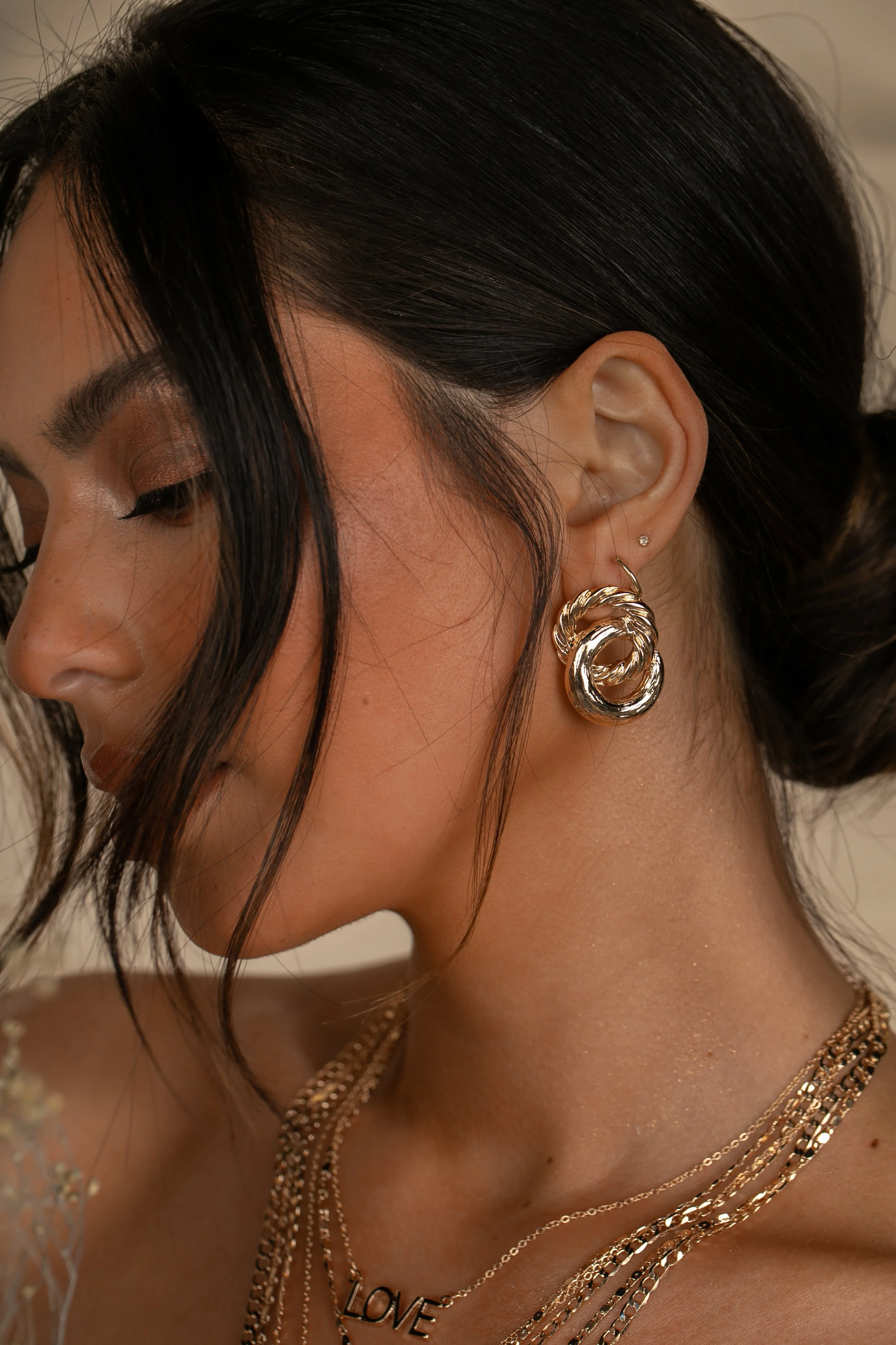 Close-up of a woman with dark hair wearing large gold hoop earrings, layered gold necklaces including one with the word 'LOVE', and a top with floral embellishments. Her eyes are closed, and her makeup is subtle.