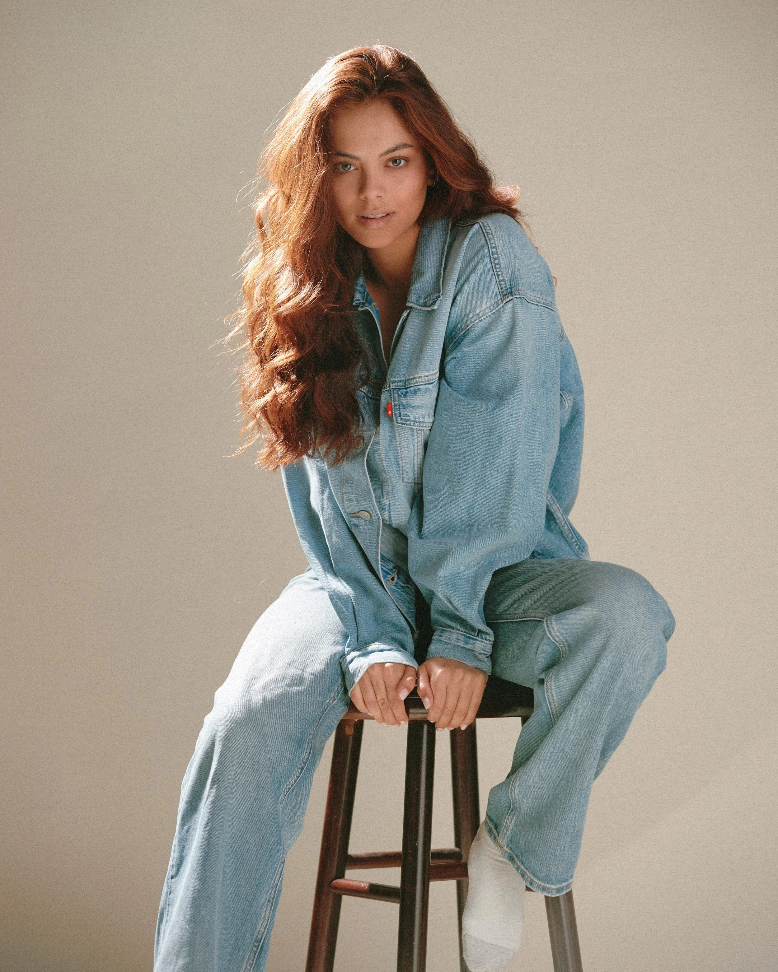 A young woman with long, wavy red hair seated on a wooden stool, wearing a denim jacket and jeans against a plain beige background.