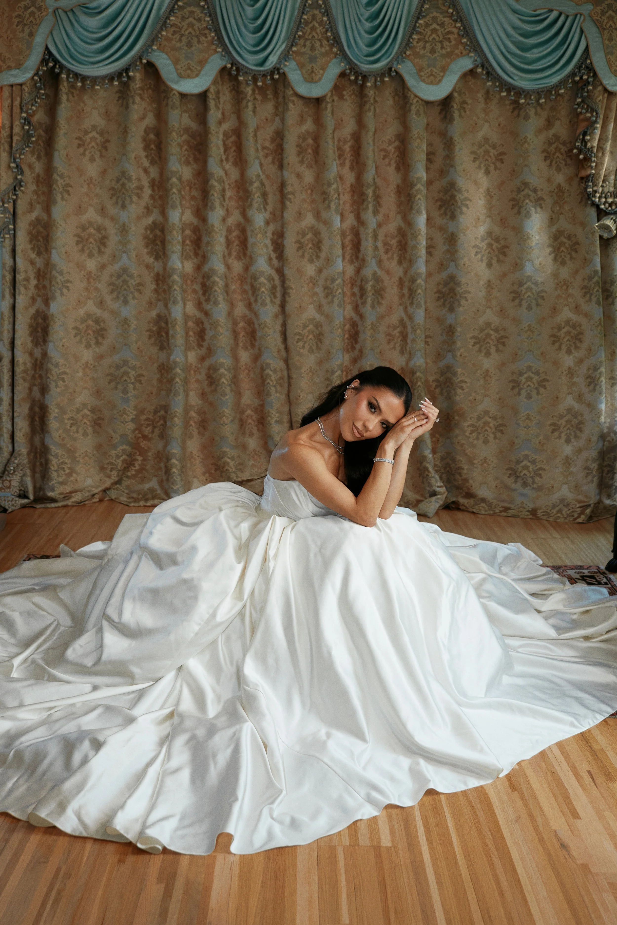 A woman in a white wedding dress sitting on the floor with her hands resting on her head against a patterned curtain backdrop.