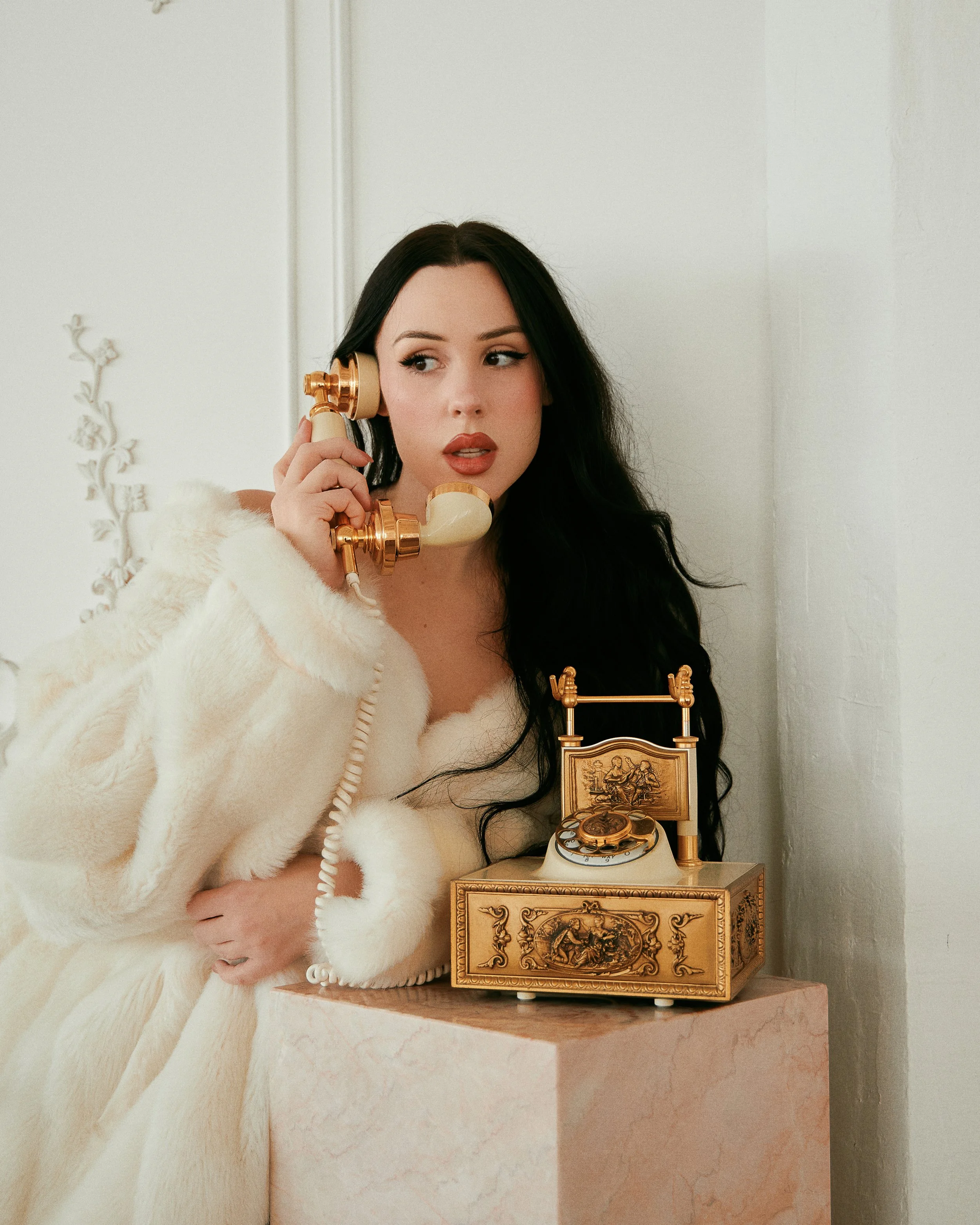 A woman with long black hair talking on a vintage rotary phone with a matching desk phone on a pink marble pedestal.