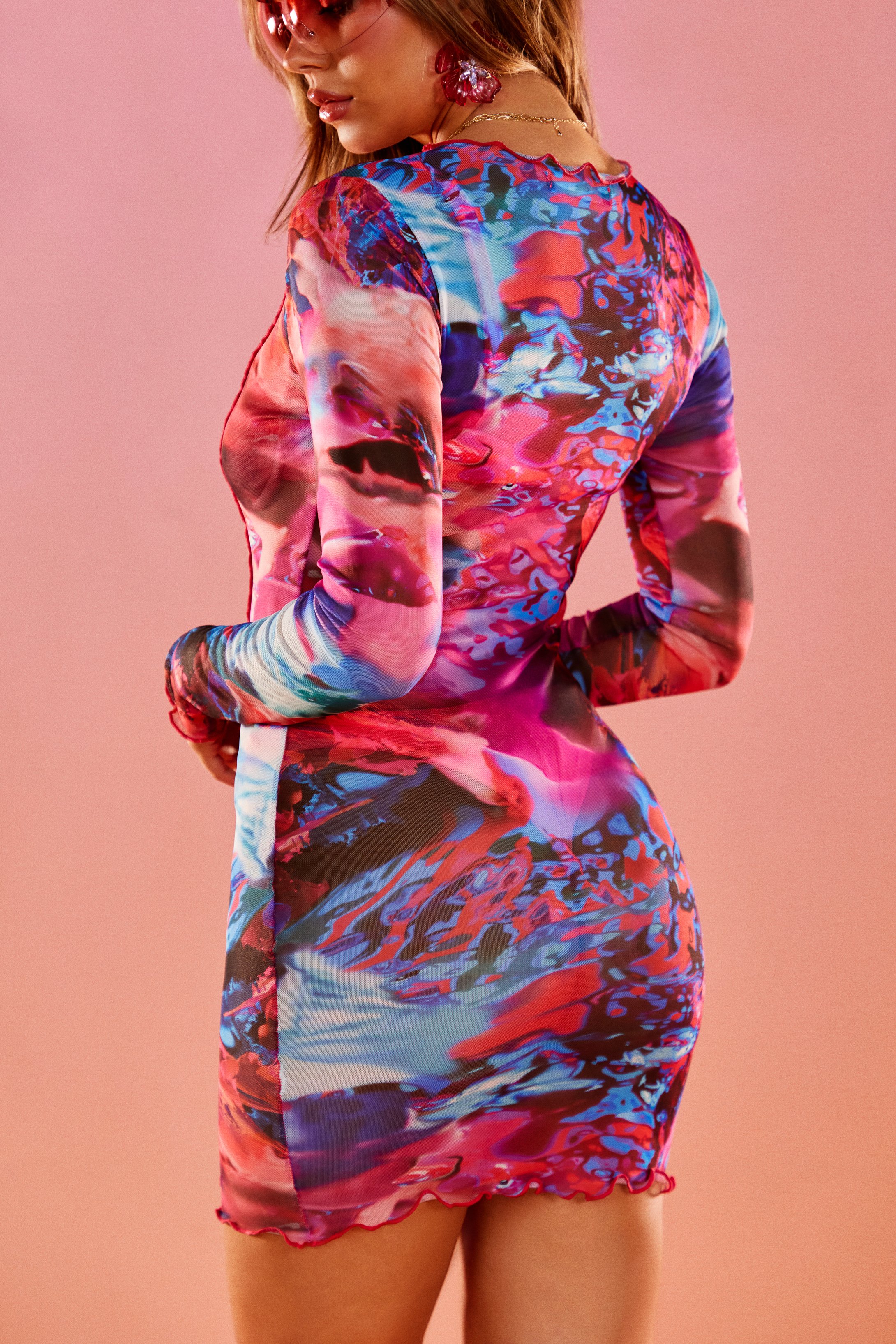 A woman in a colorful, abstract-patterned dress standing with her back to the camera against a pink background.