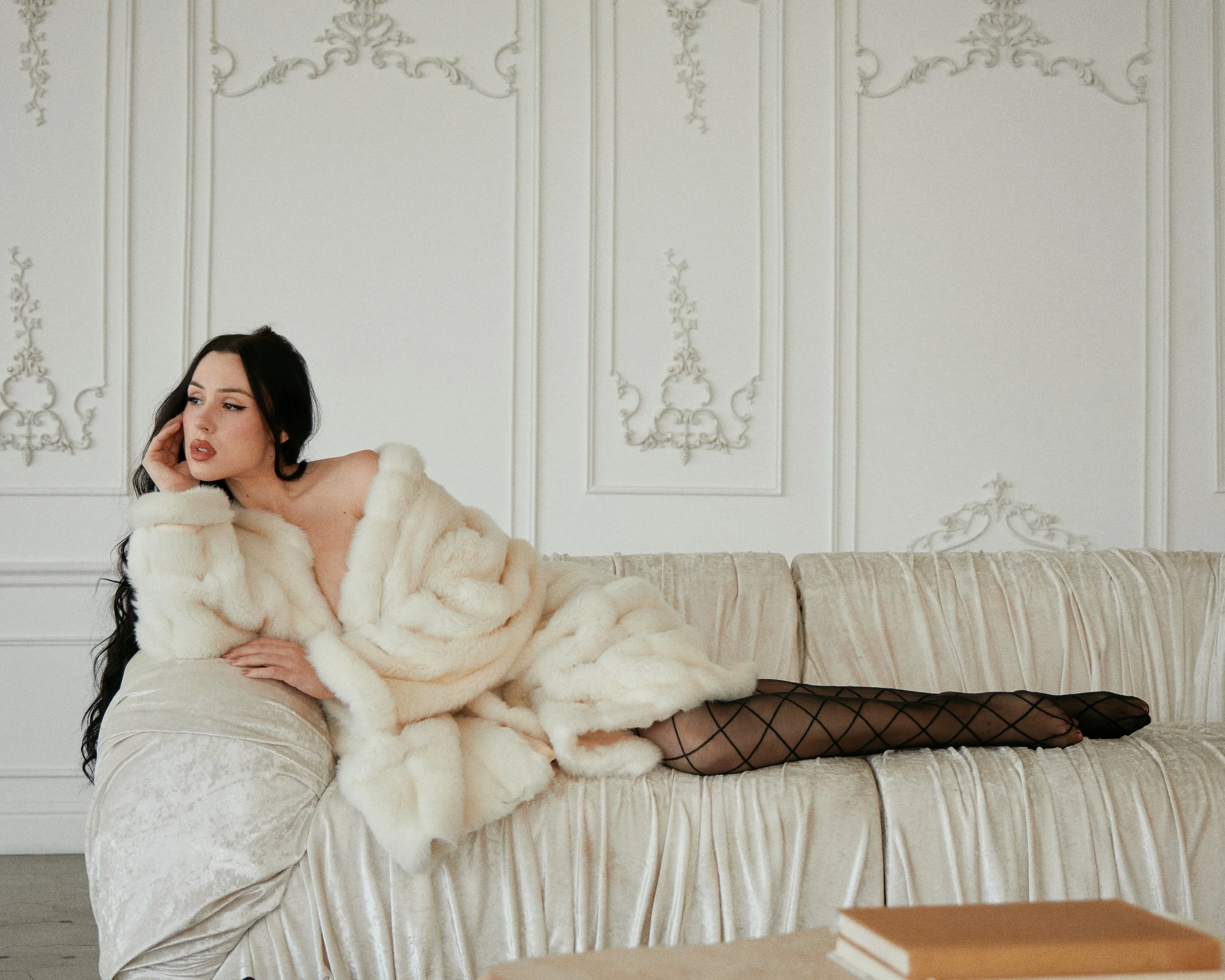 A woman with dark hair lying on a vintage cream-colored velvet sofa, wearing black fishnet stockings and a luxurious cream-colored fur coat, in a room with ornate white wall paneling.