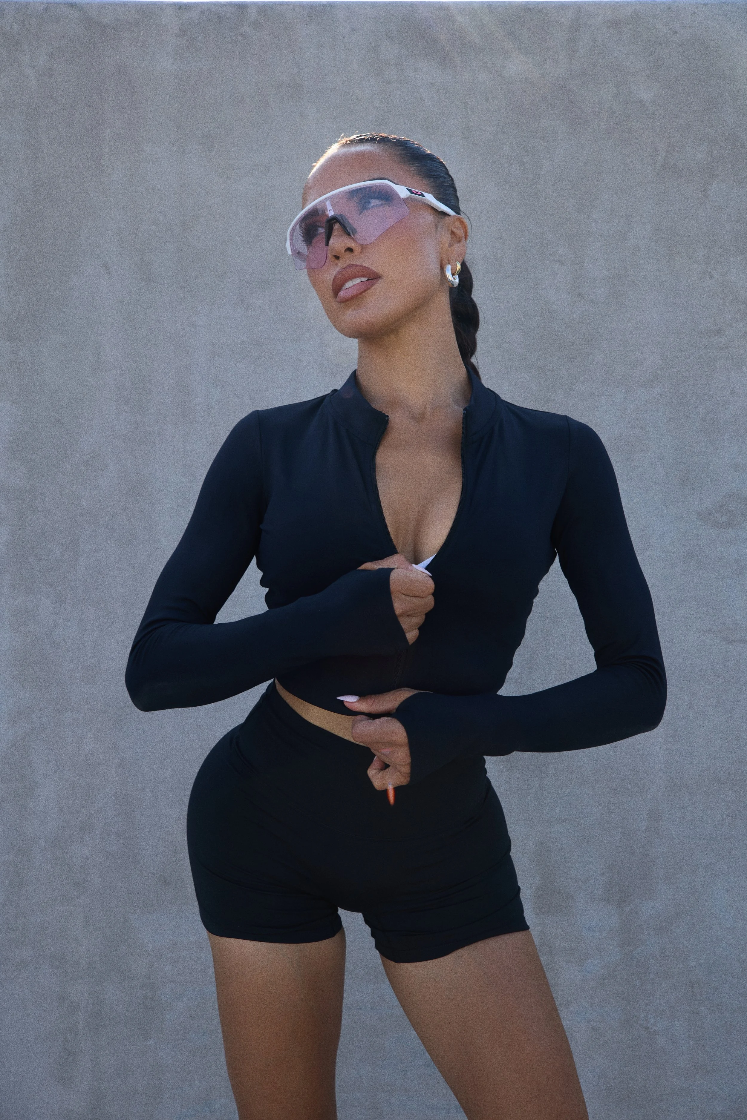 A woman wearing sporty workout clothes, large wrap-around sunglasses, and hoop earrings, standing against a plain wall, with her hair in a braid.