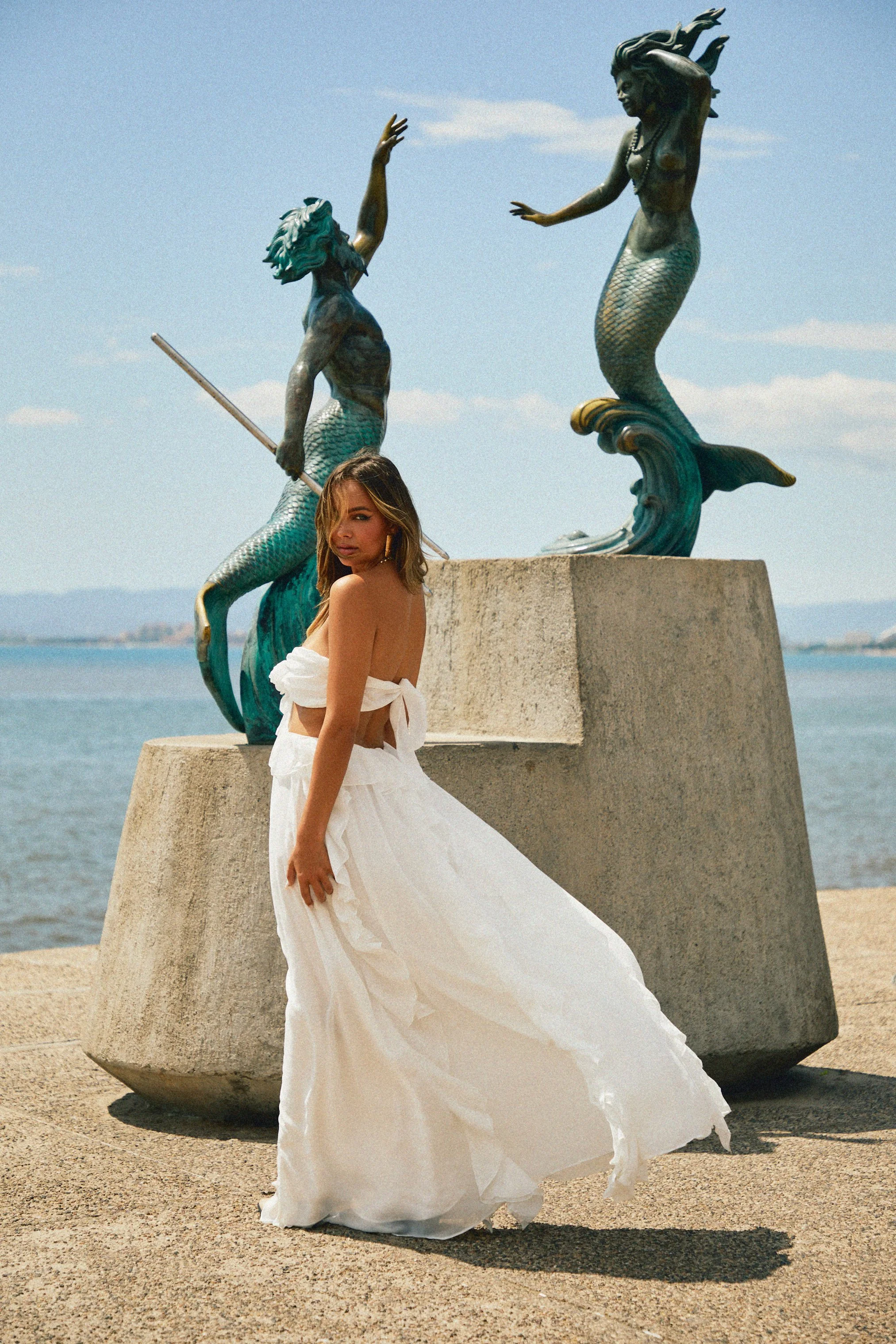 A woman in a white flowing dress standing near ocean with two blue-green mermaid statues behind her.