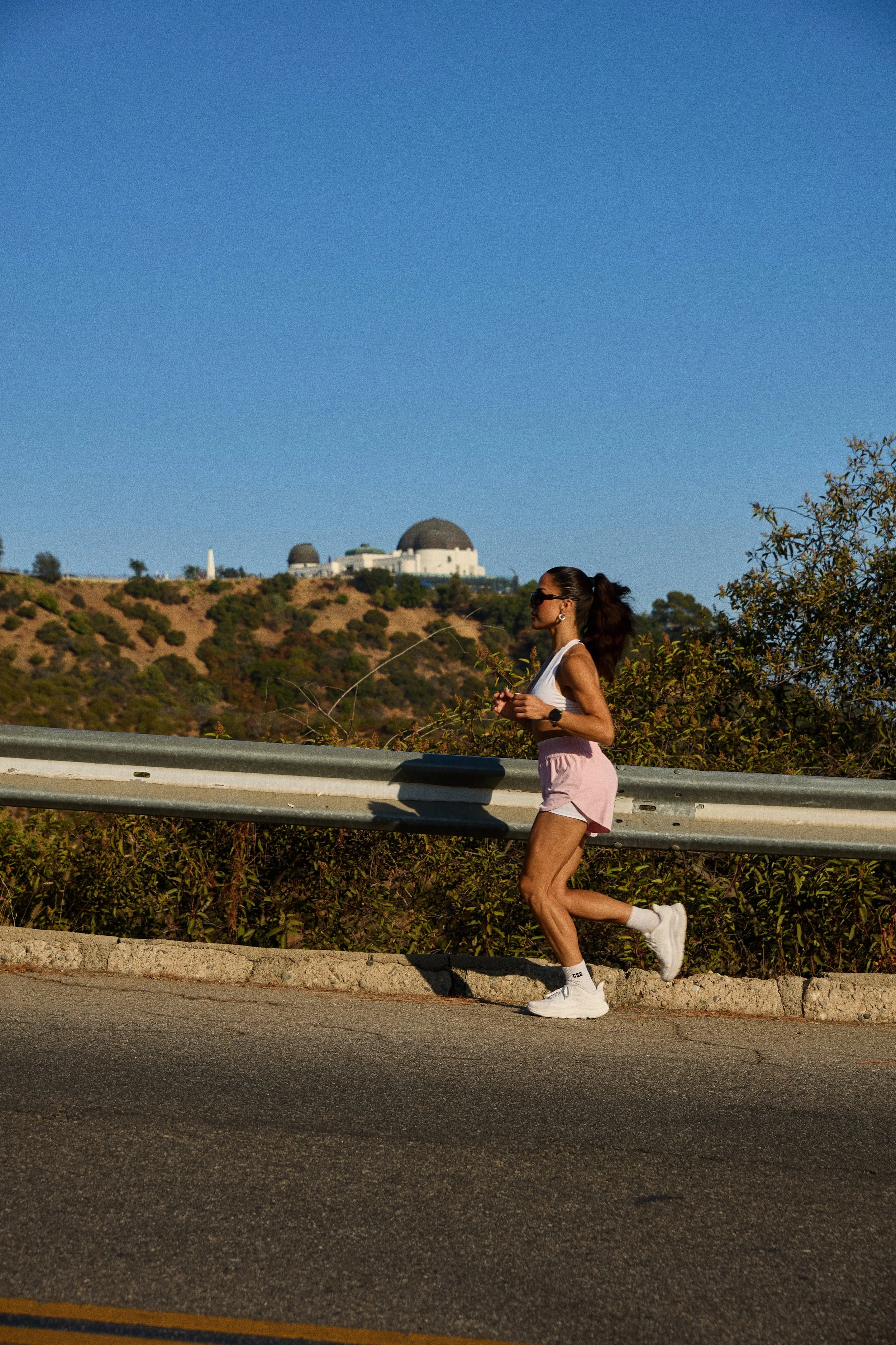 A woman jogging along a roadside with the Griffith Observatory visible in the background, under a clear blue sky.
