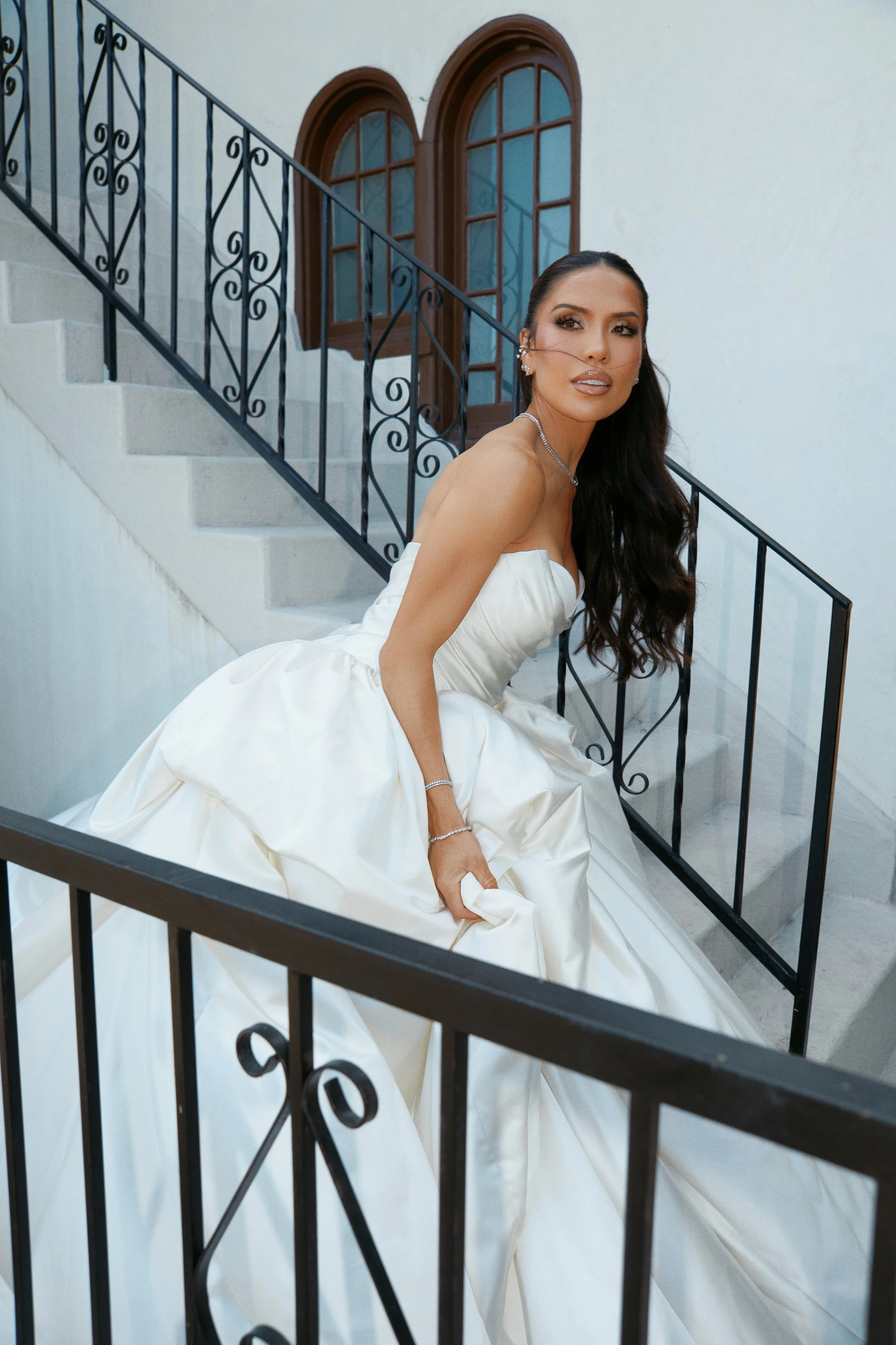 A woman in a white wedding dress sitting on a staircase with black wrought iron railing, looking at the camera with dark, wavy hair and jewelry, in front of arched wooden windows on a white wall.