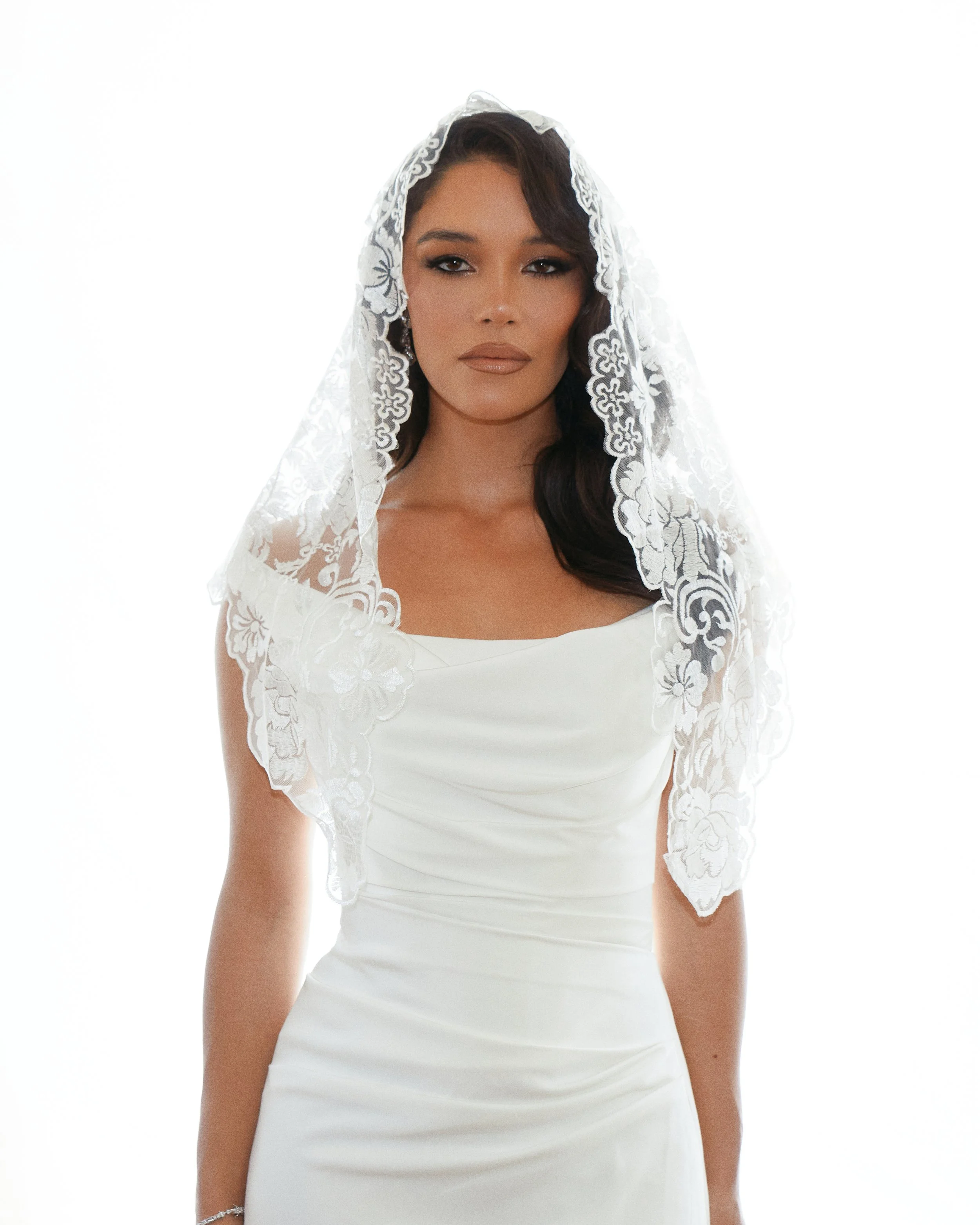 A woman in a wedding dress with a lace veil over her head, standing against a white background.