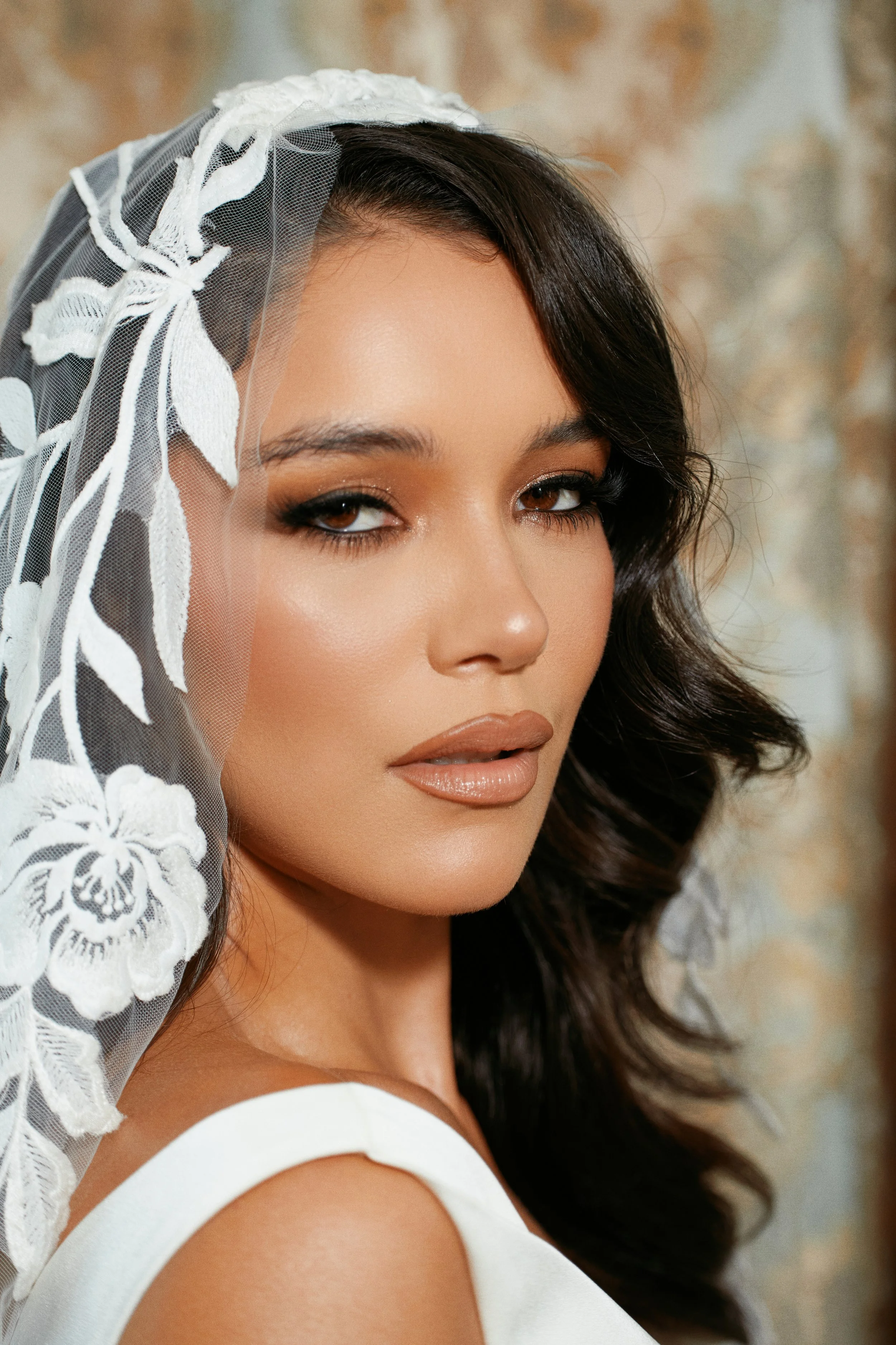 Close-up of a woman with dark wavy hair and light makeup, wearing a white dress and a lace veil with floral embroidery.