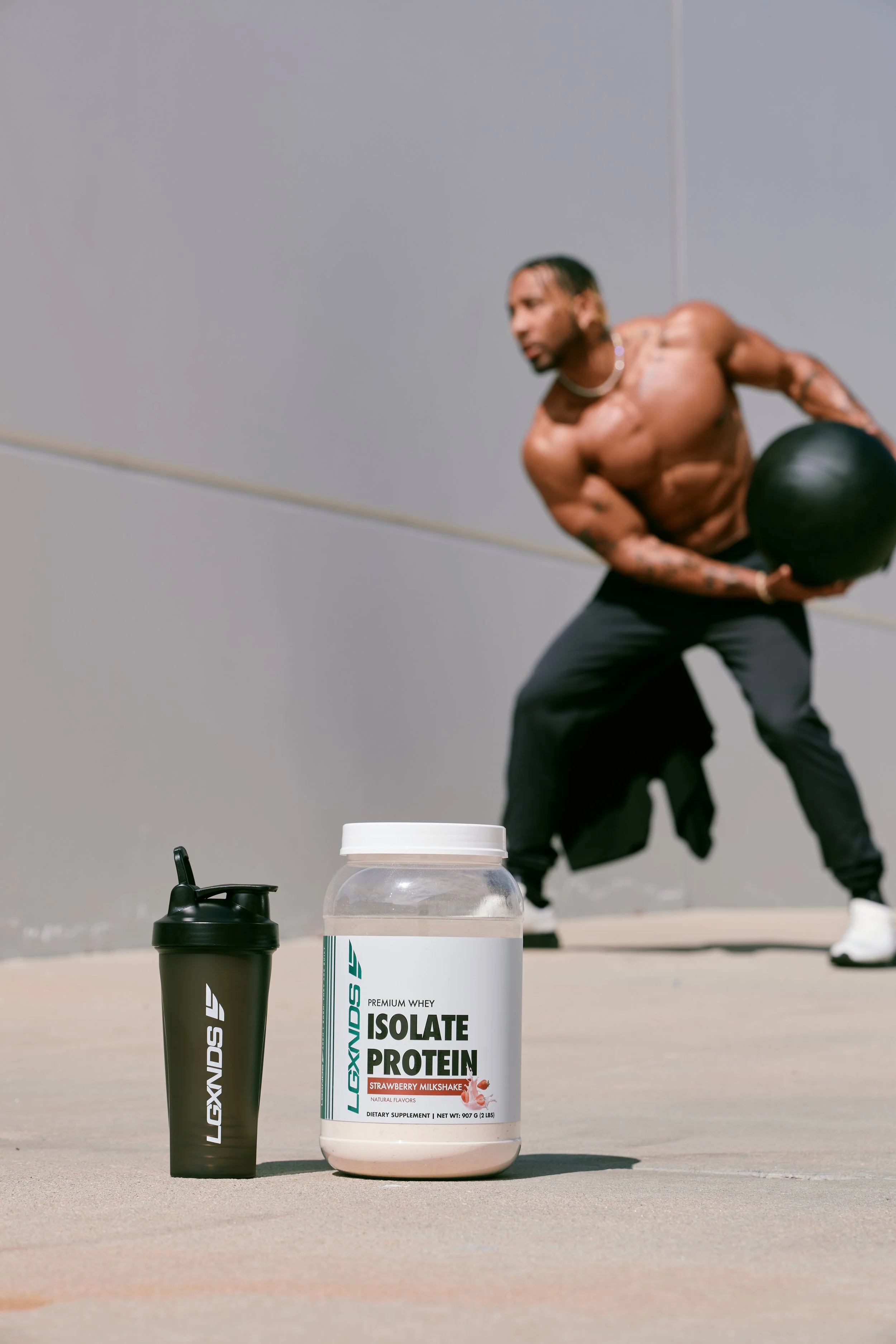A container of Legionds isolate protein powder and a shaker bottle with Legionds logo placed on the floor, with a muscular man in the background holding a medicine ball during a workout.