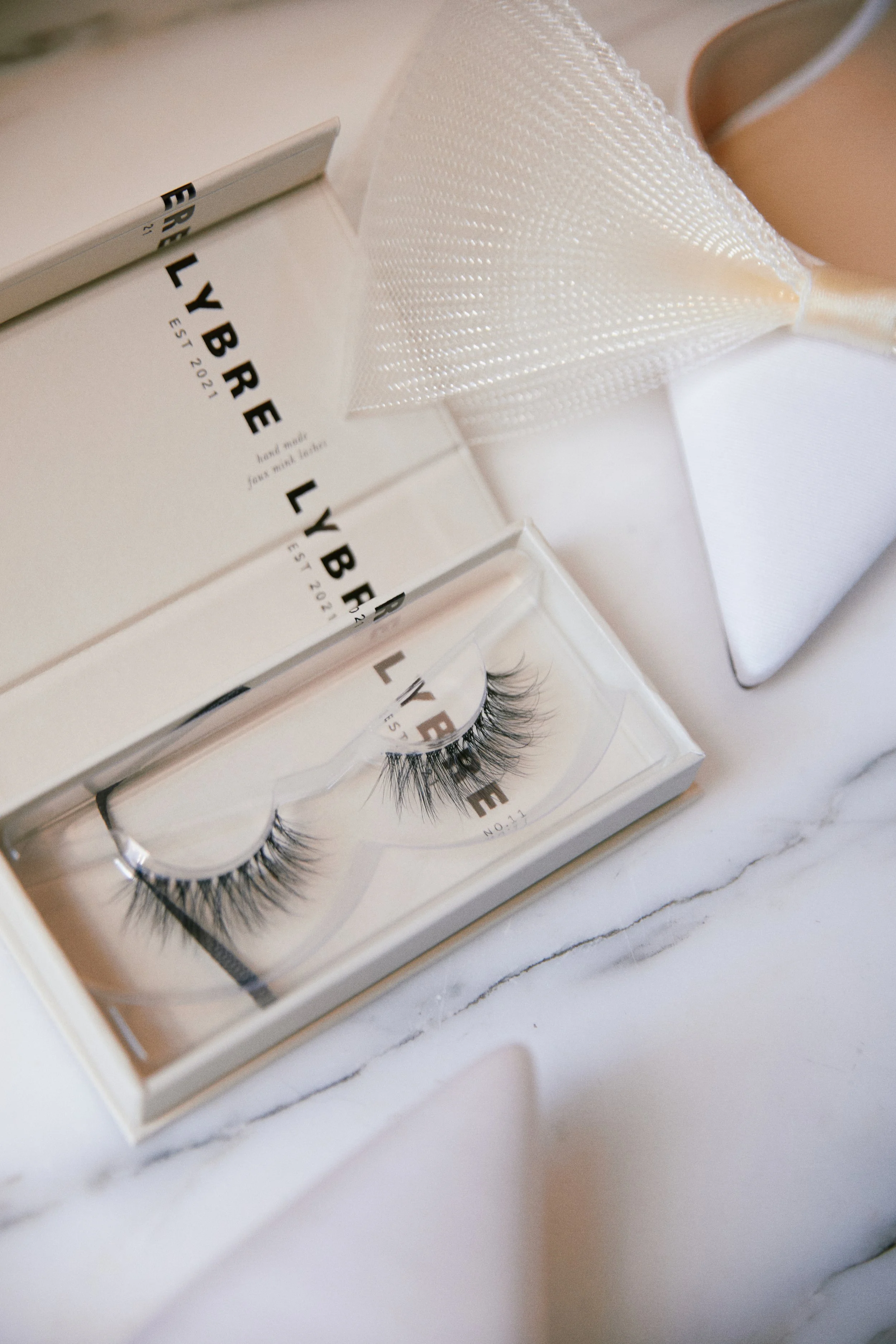 A pair of false eyelashes in a clear plastic case, with a white box labeled 'LYBE' and a textured white applicator or cover, all placed on a white marble surface.