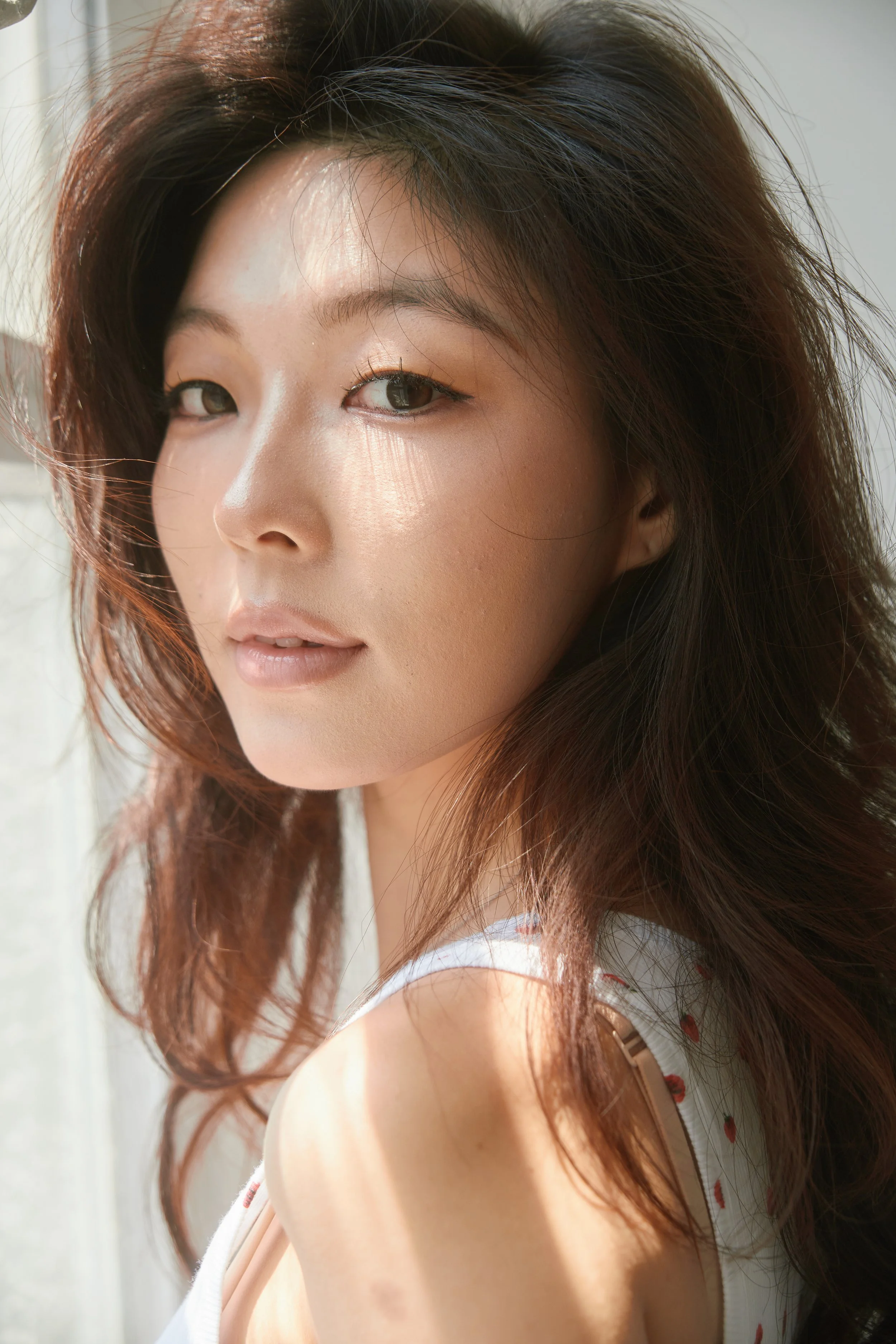Close-up of a woman with wavy, reddish-brown hair, looking at the camera with a subtle smile, indoors with natural light.