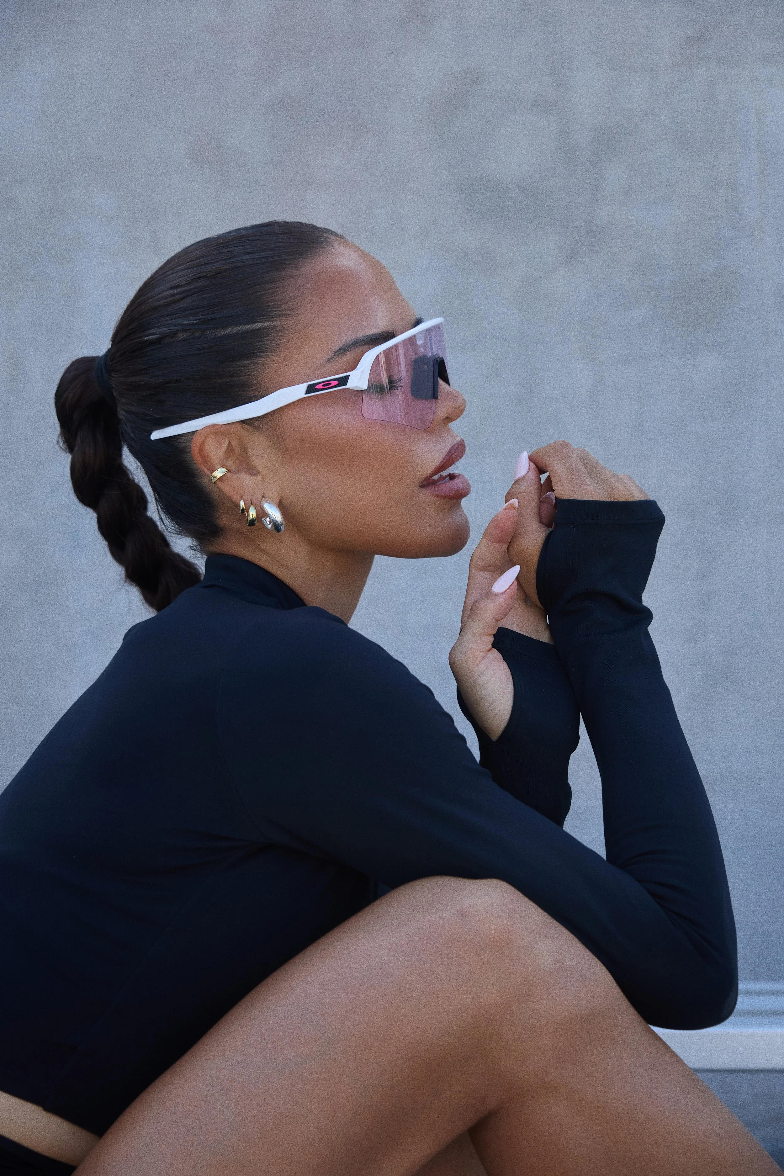 A woman with braided hair, multiple earrings, and wearing large sunglasses, a black long-sleeve top, and gloves is sitting against a plain grey wall while looking to the side.