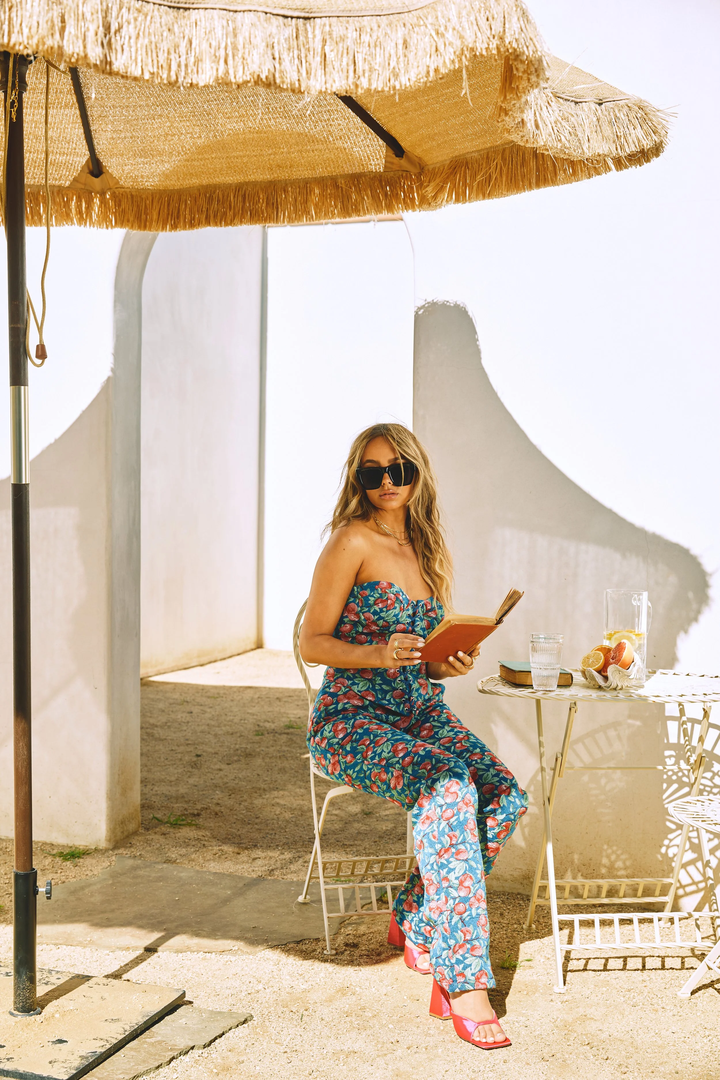 A woman sitting at an outdoor table reading a book under a straw umbrella on a sunny day.