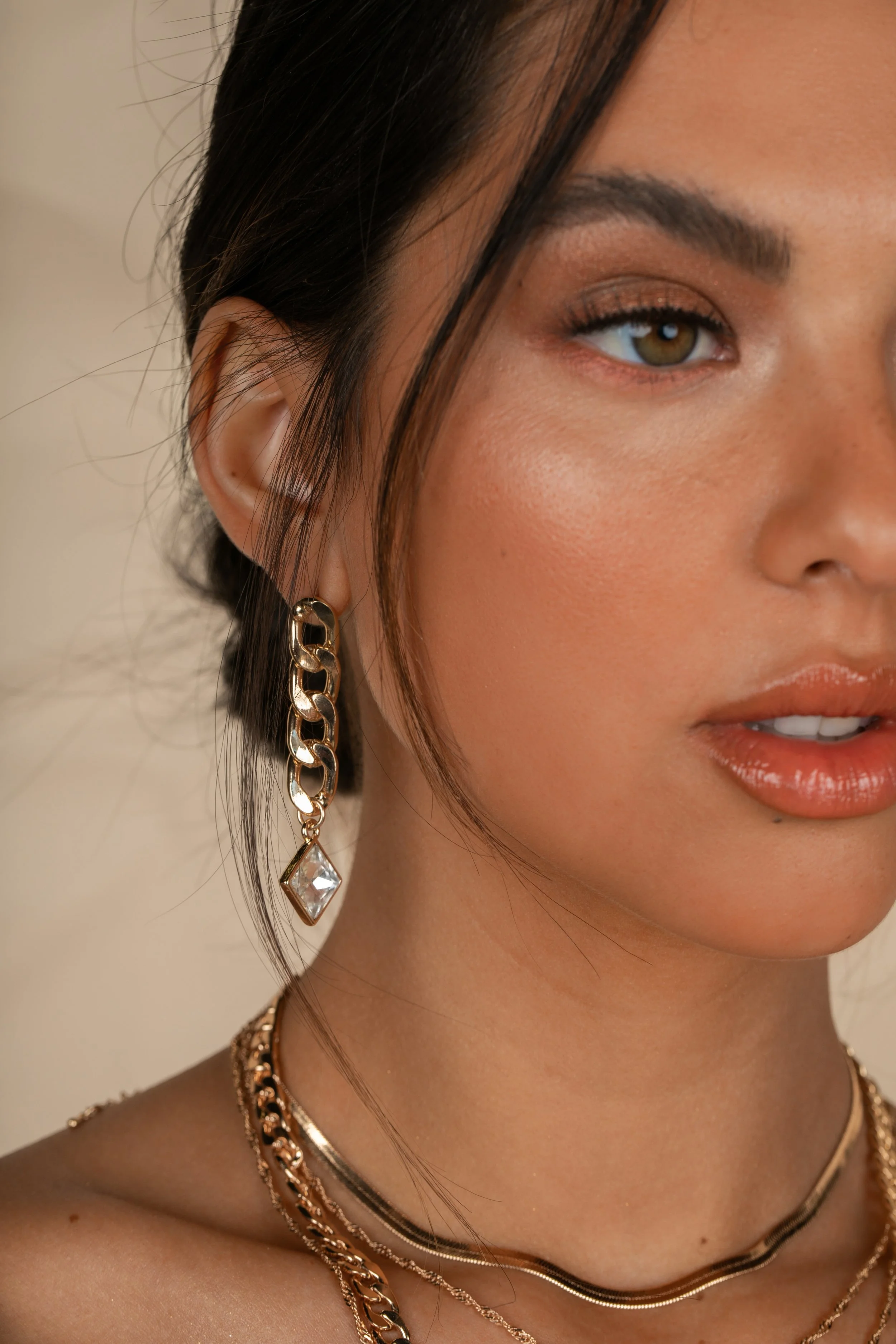 Close-up of a woman with styled dark hair, wearing gold jewelry including a chain link earring with a diamond-shaped gemstone, layered necklaces, and makeup with natural tones.