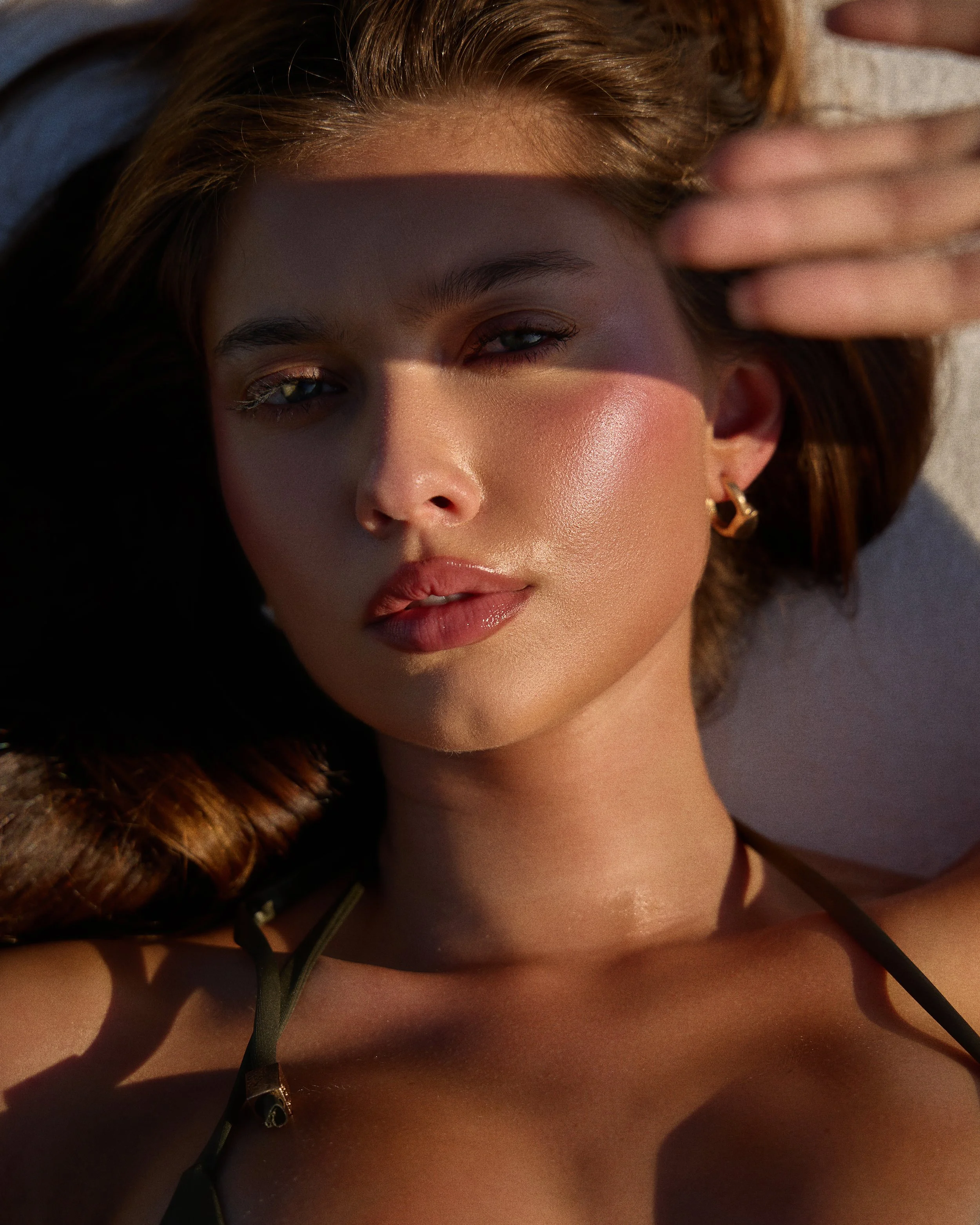 Close-up of a young woman with light skin, brown hair, and hoop earrings lying on her back on the beach. She is shielding her eyes from the sun with her hand, and sunlight is casting a shadow across her face.