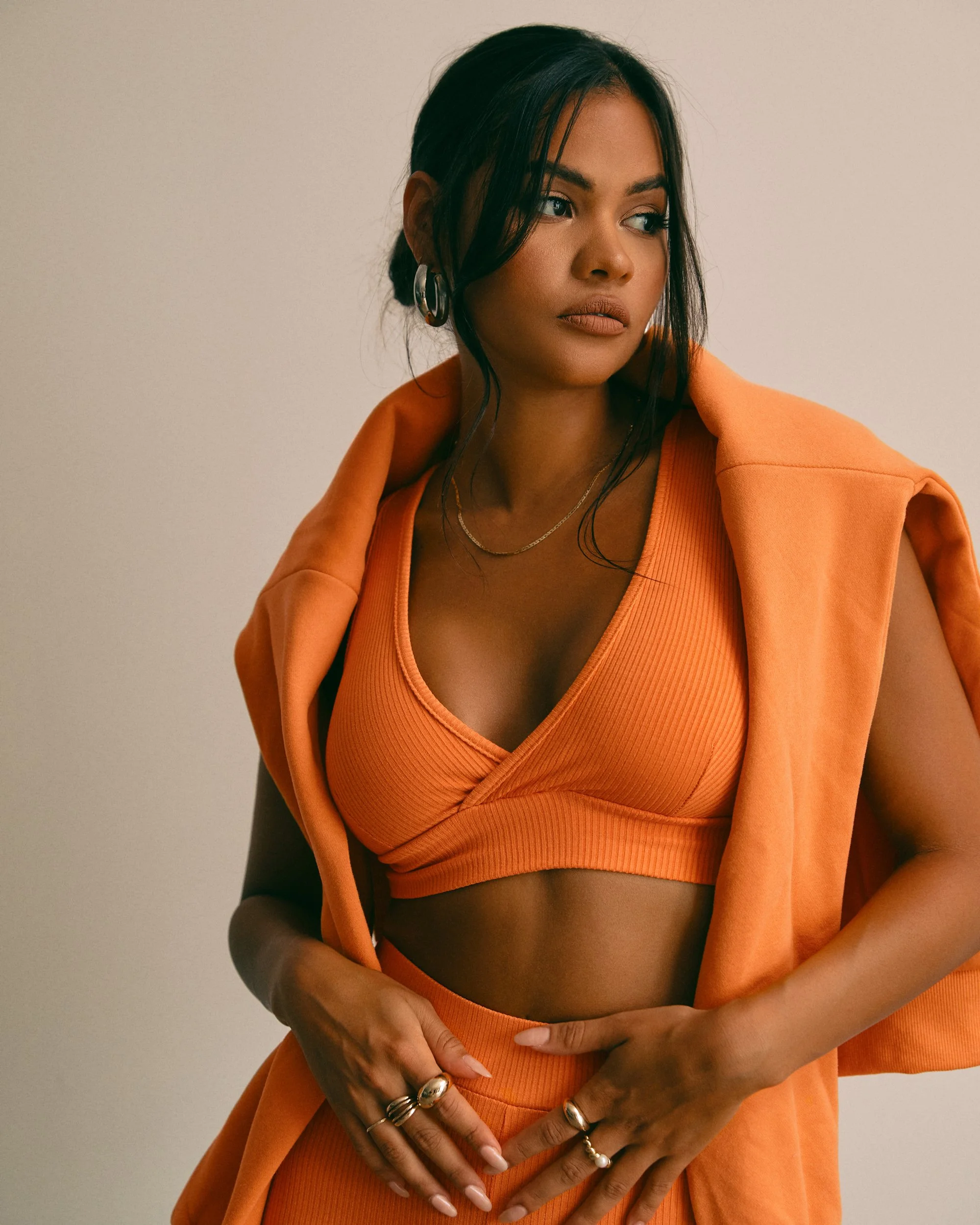 A woman wearing an orange sports bra and matching pants, with an orange blazer draped over her shoulders, posing against a neutral background.