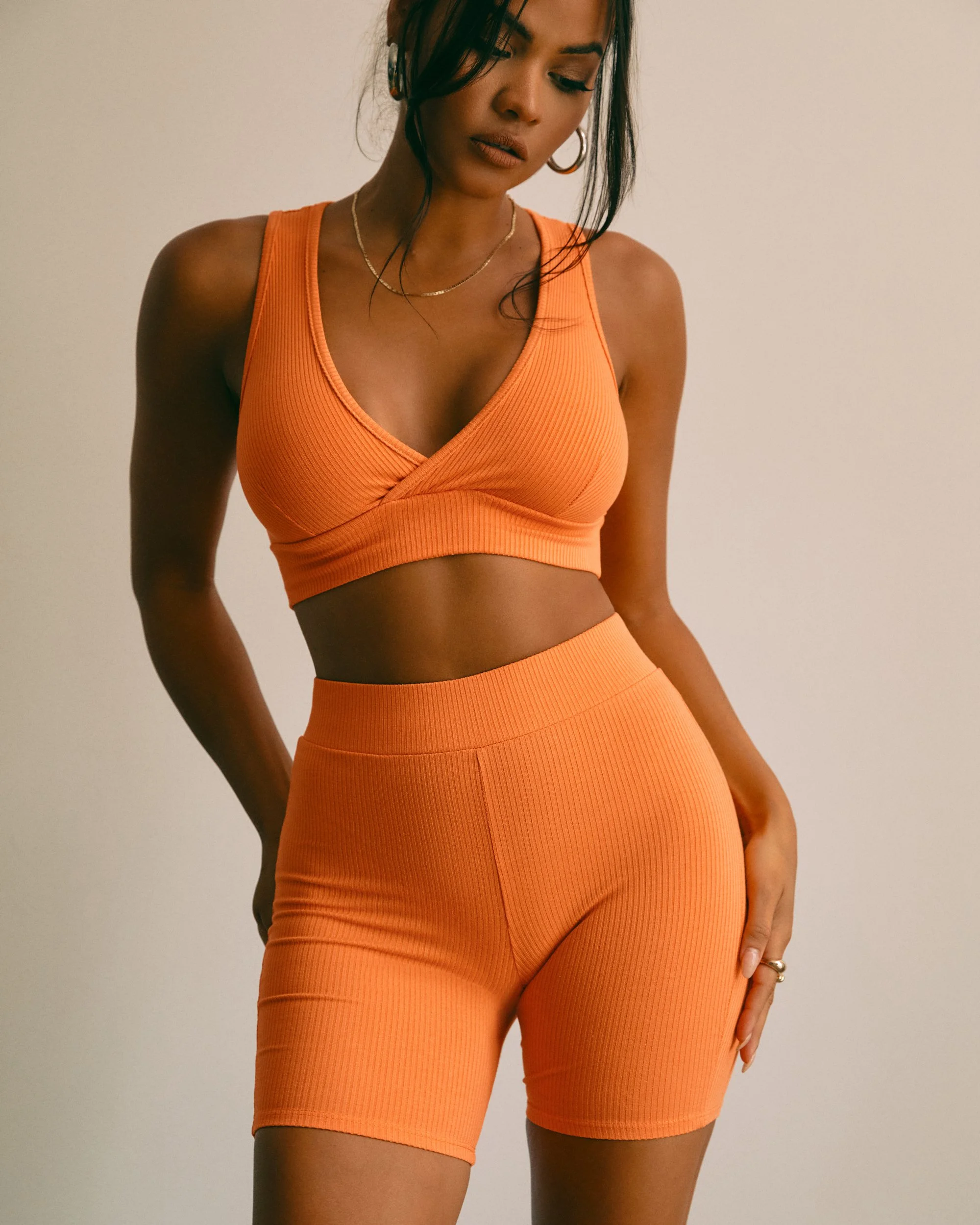 A woman wearing an orange workout set with a crop top and matching shorts, standing against a plain wall.