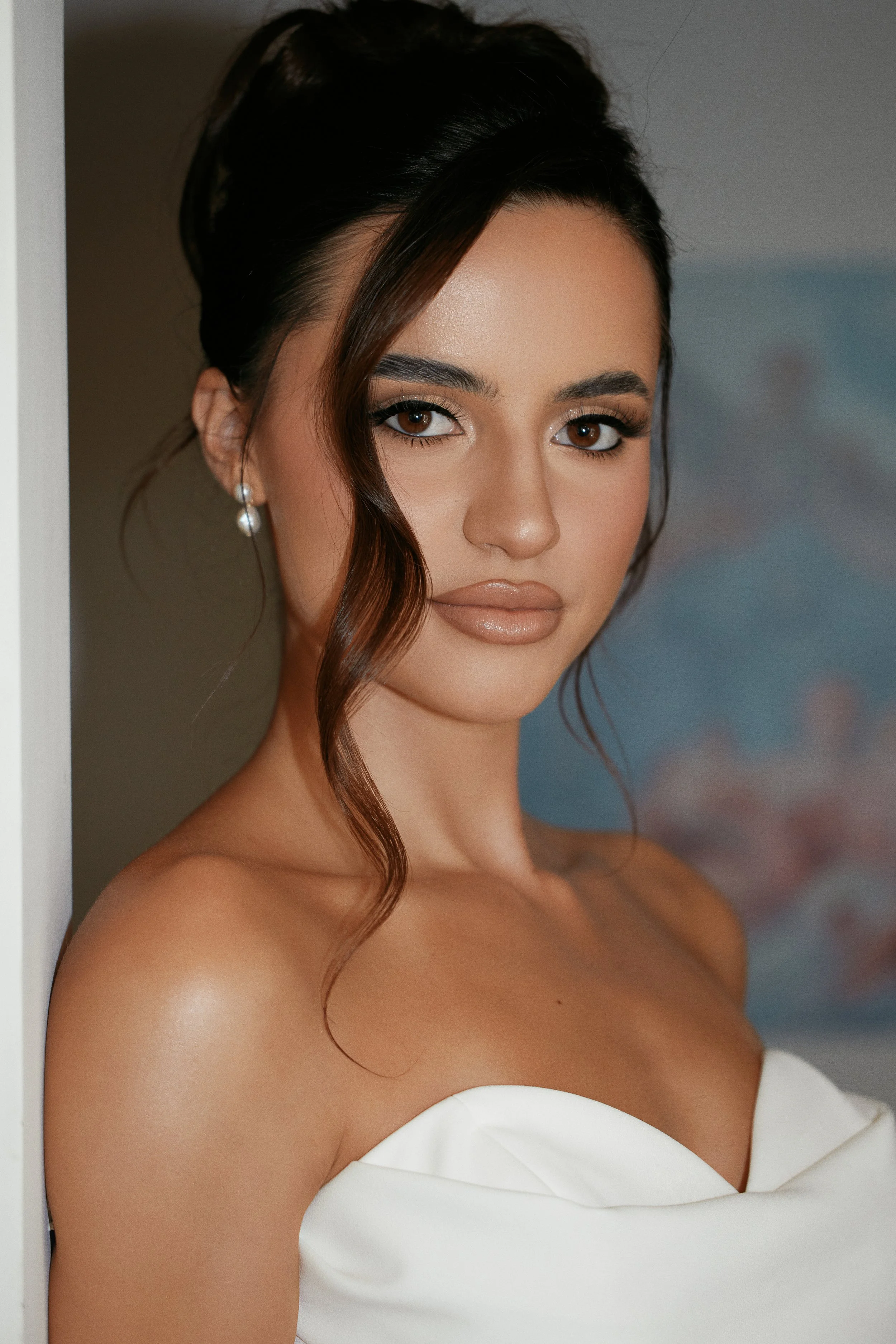 A woman with dark hair styled in an elegant updo with a loose, wavy strand, wearing pearl earrings and a strapless white dress, standing against a neutral background.