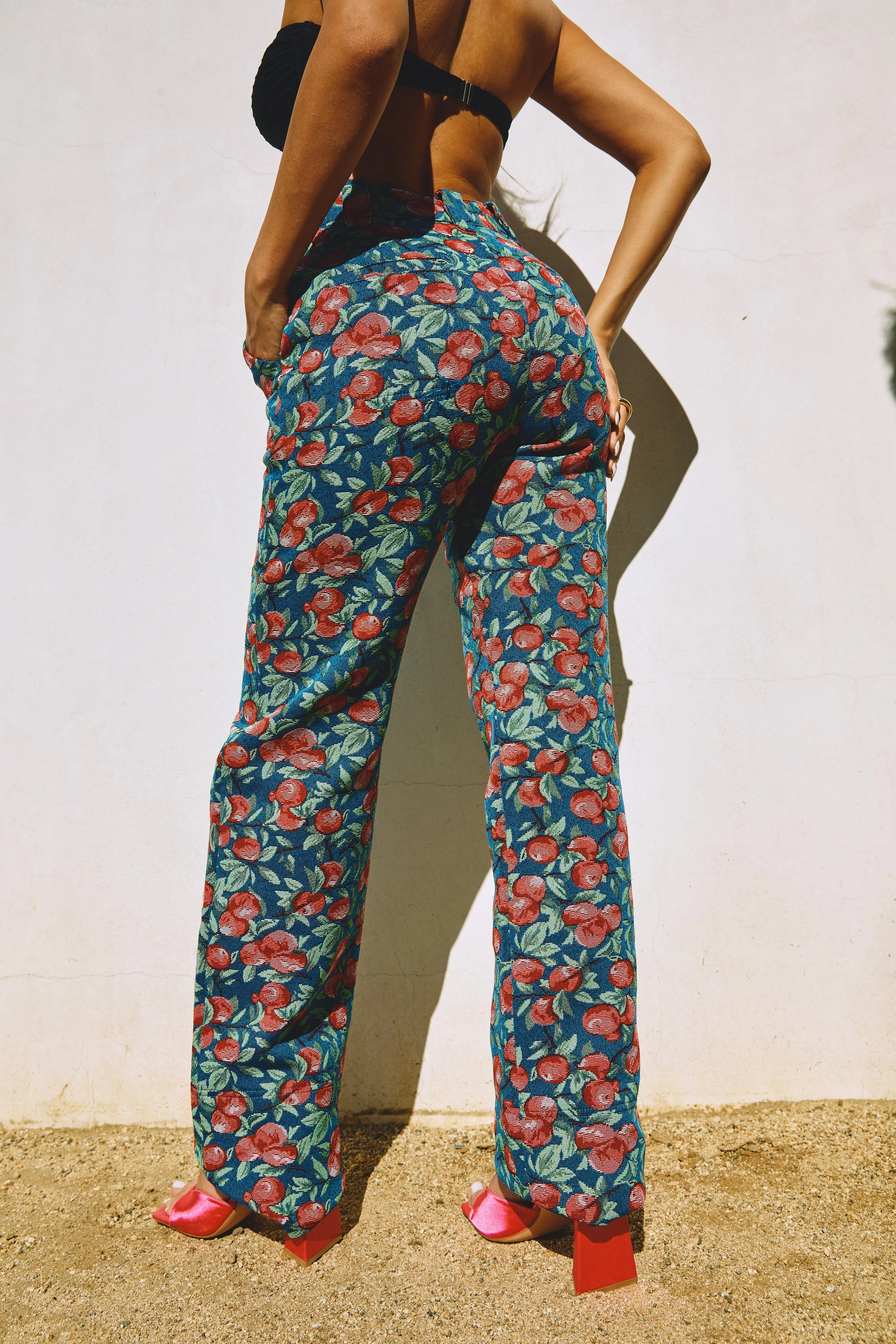 Woman wearing floral pants and pink high heels standing against a white wall, viewed from the back.
