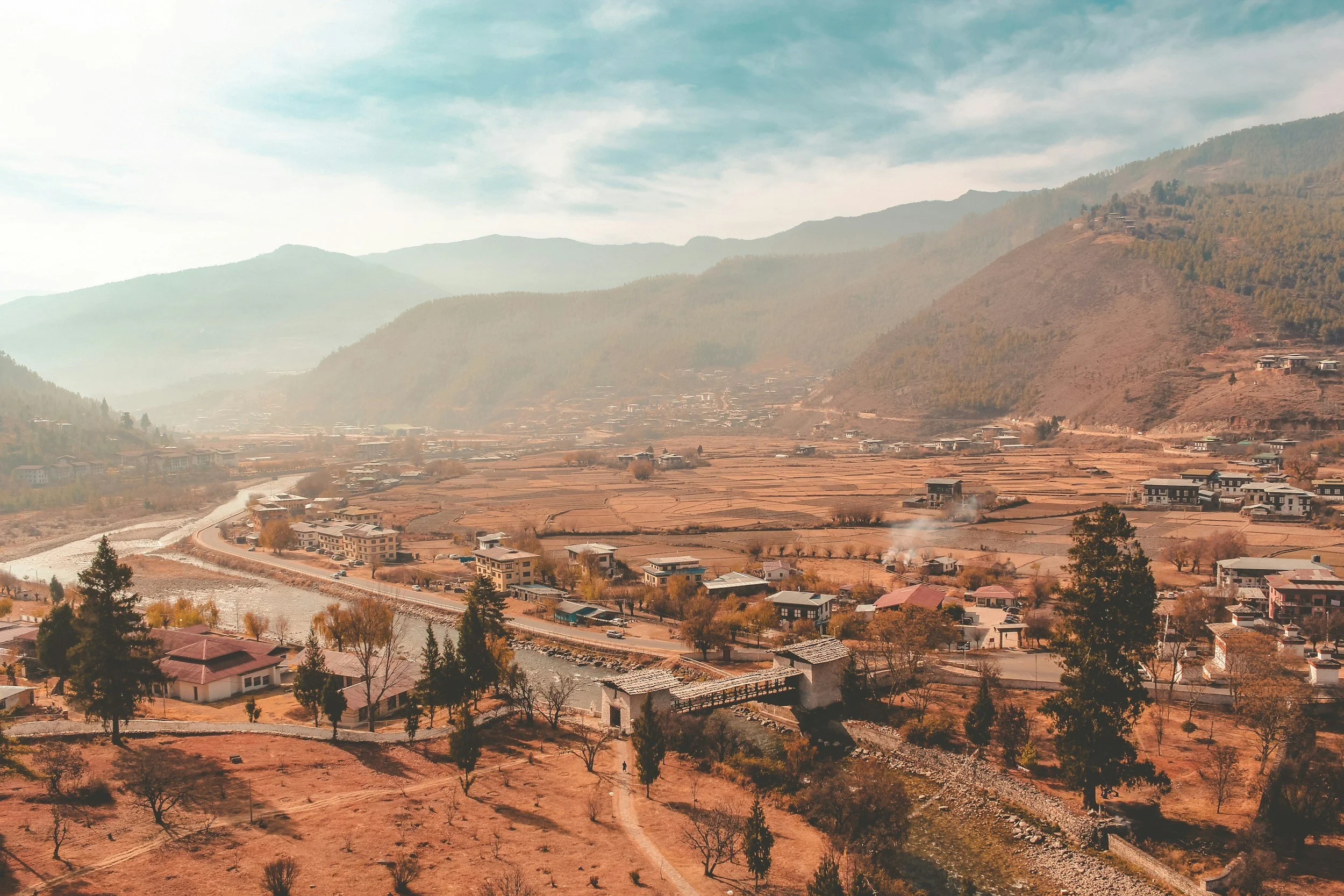 A small town in a valley surrounded by mountains, with a river running through it, houses, trees, and smoke rising from some buildings.