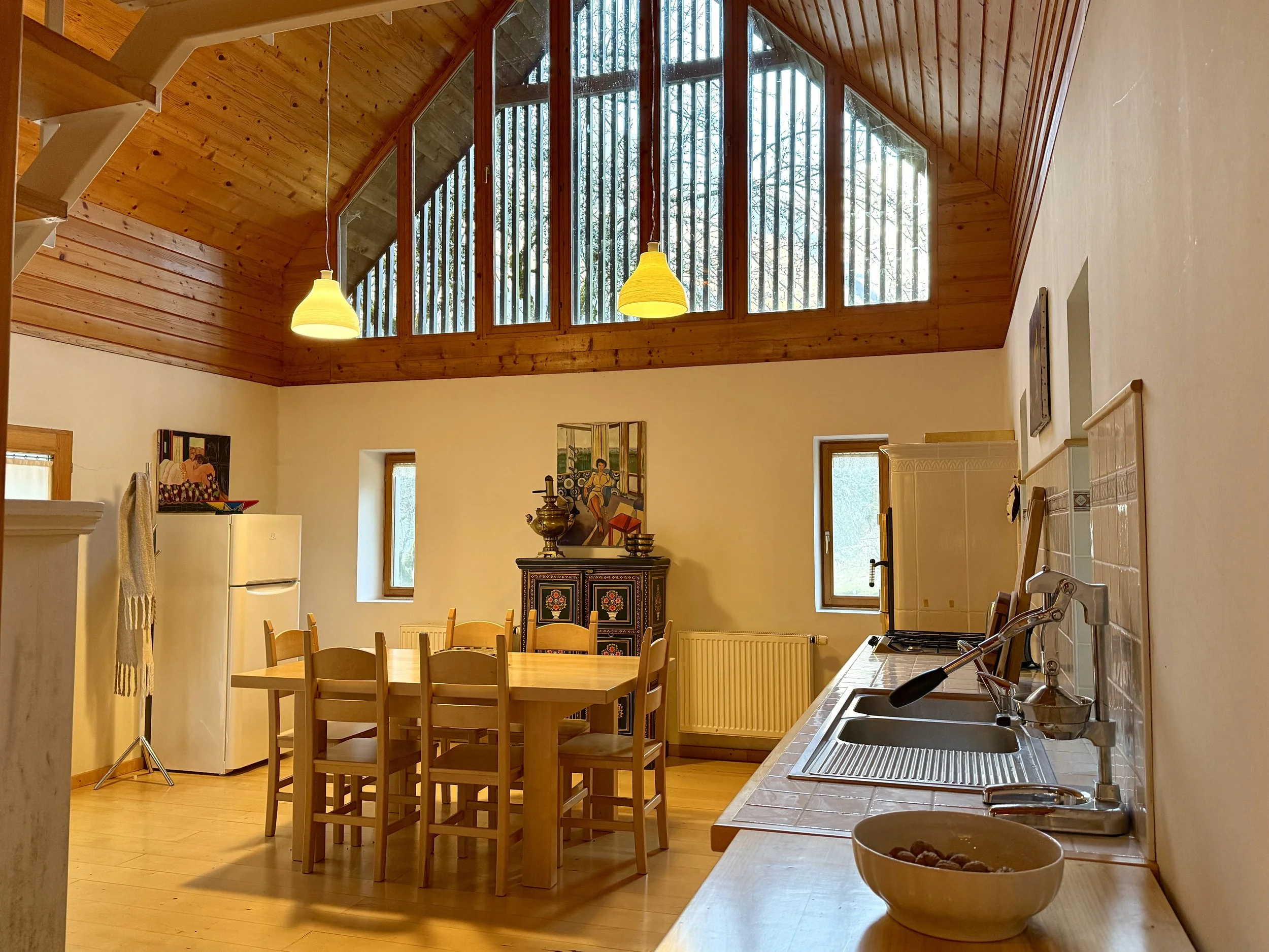 A cozy kitchen with a wooden vaulted ceiling and large windows letting in natural light. There is a kitchen counter with a sink and a bowl of walnuts, a dining table with multiple chairs, a refrigerator, and decorative artwork on the wall.