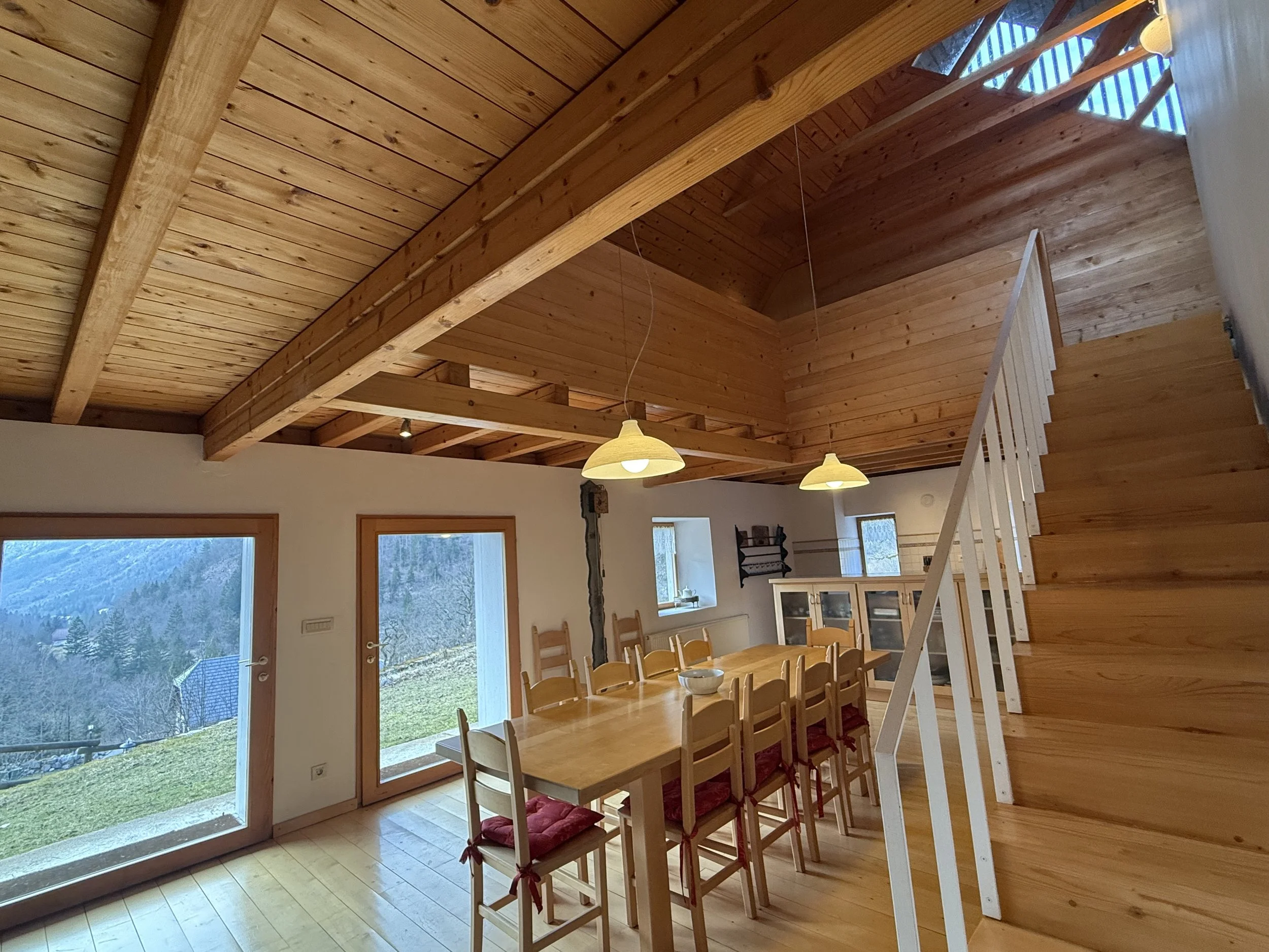 Interior view of a wooden house with a dining area, large glass doors, and windows showing mountain scenery outside; wooden staircase leading to upper level, wooden beams and ceiling, and pendant lights hanging above the dining table.