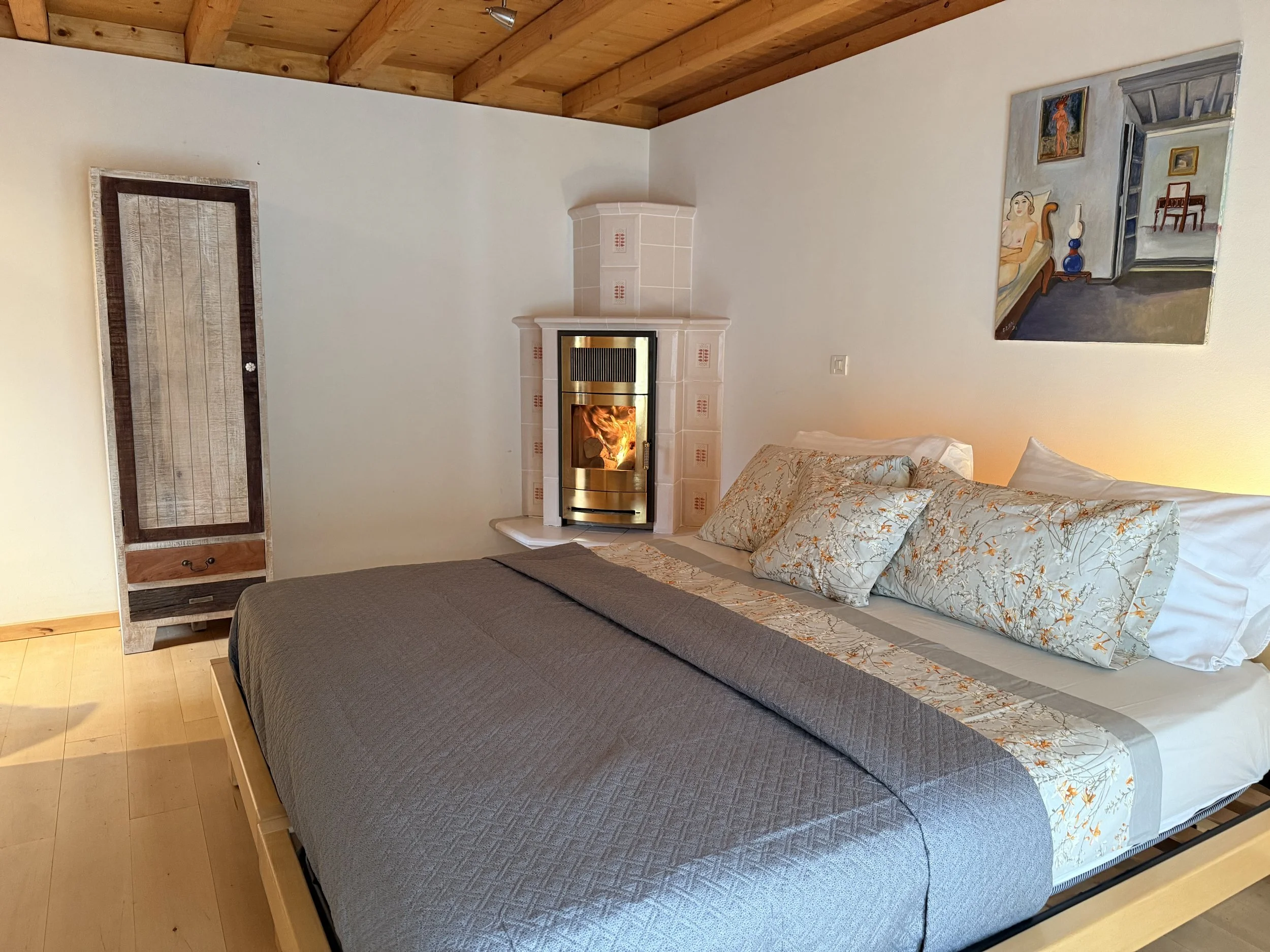 Cozy bedroom with a large bed featuring a gray quilt and floral pillows, a corner fireplace with a fire, wooden ceiling beams, and a colorful artwork on the wall.