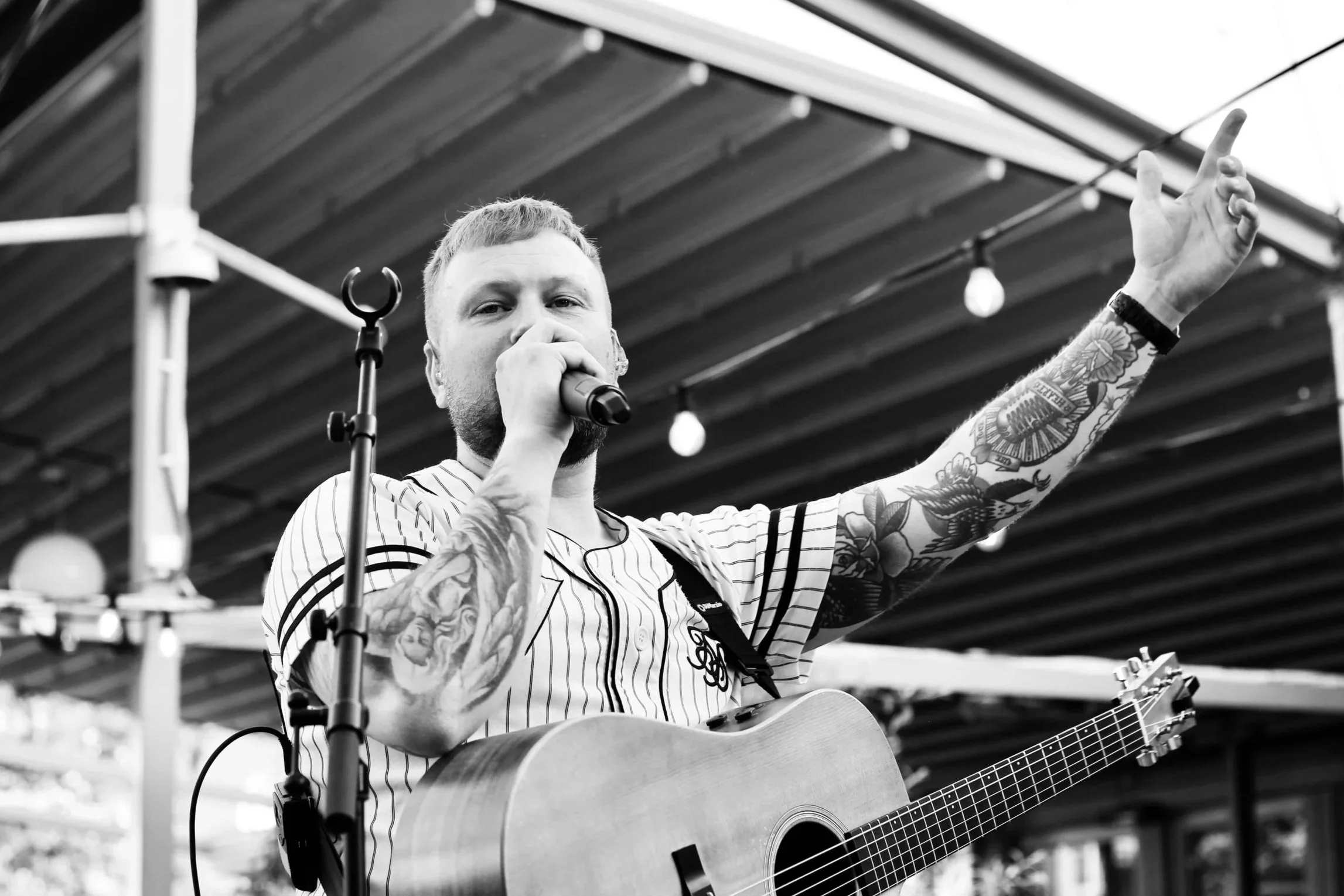 Manlig sångare sjunger i mikrofon på scen, bär vit- och svartrandig t-shirt. Har tatueringar på armarna och en gitarr runt halsen.