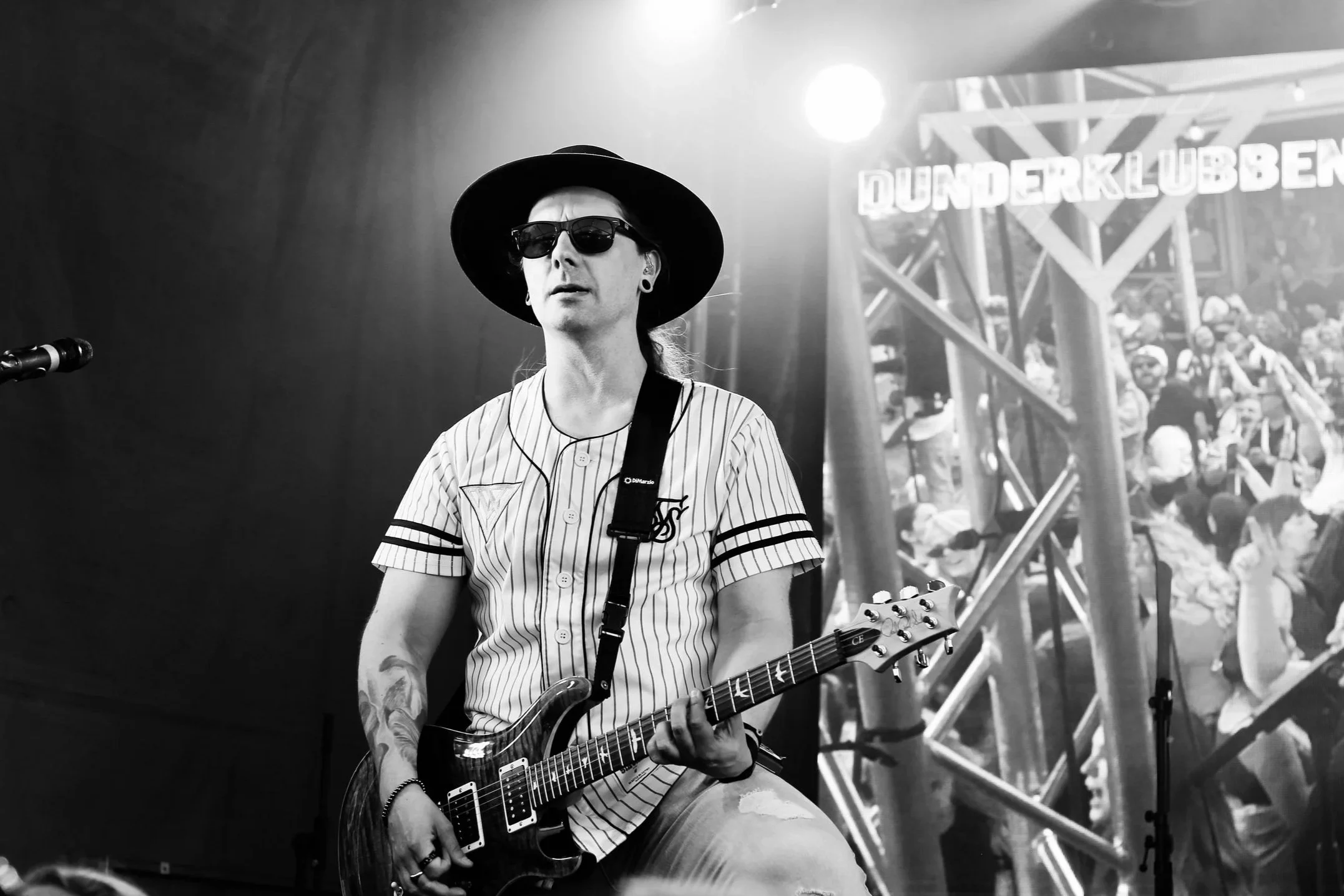 Manlig gitarrist står på scenen och spelar i en vit- och svartrandig t-shirt, solglasögon och svart hatt. I bakgrunden syns en bildskärm på ett publikhav.