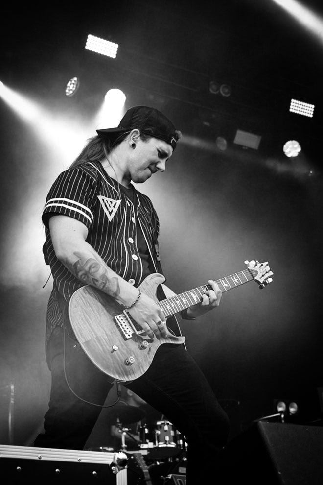 Gitarrist från Dunderklubben coverband spelar live på scen, iförd svart randig tröja och keps bakåtvänd, omgiven av scenljus och rökeffekter.