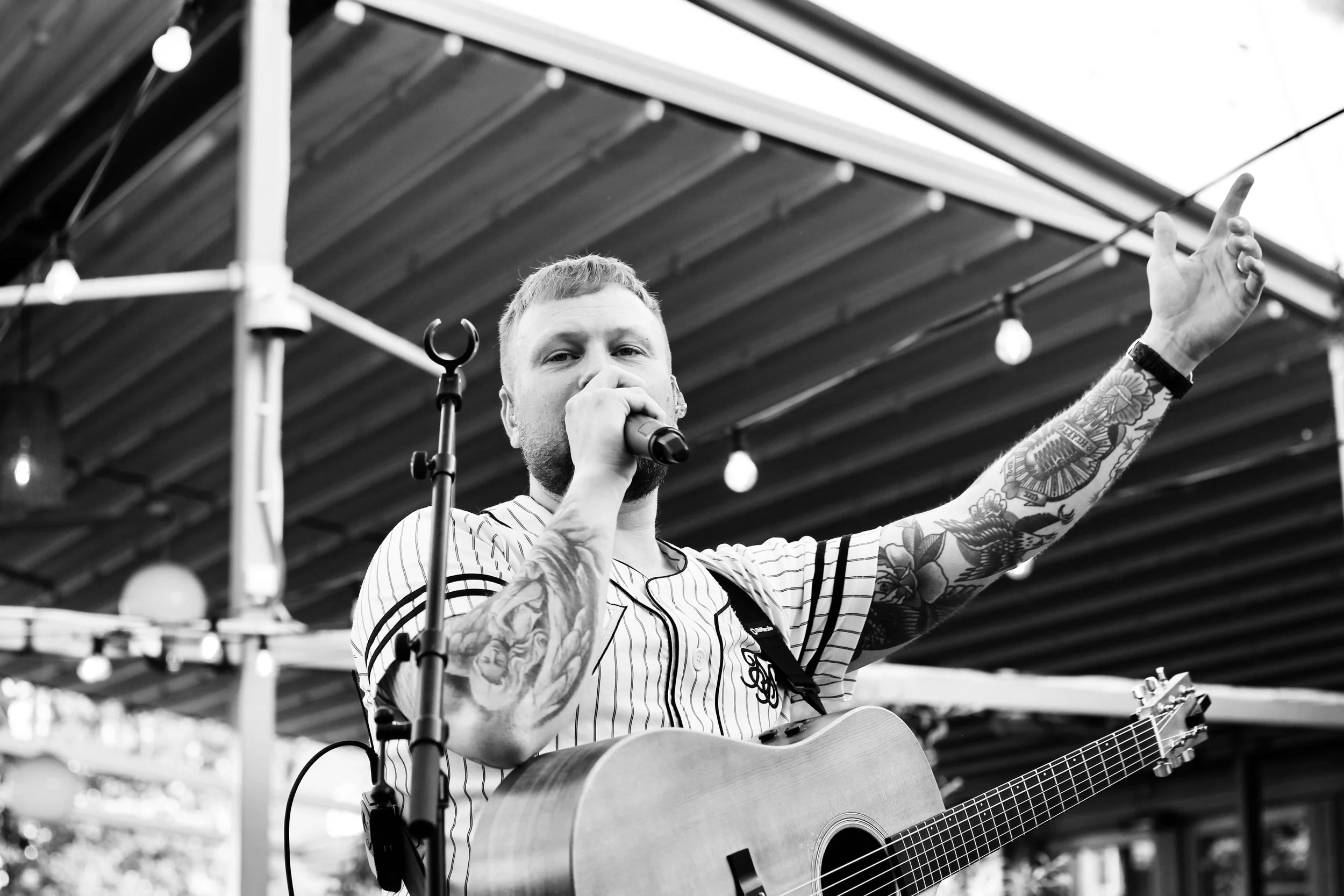 Tatuerad manlig kortklippt sångare sjunger i mikrofon med en akustisk gitarr runt halsen..