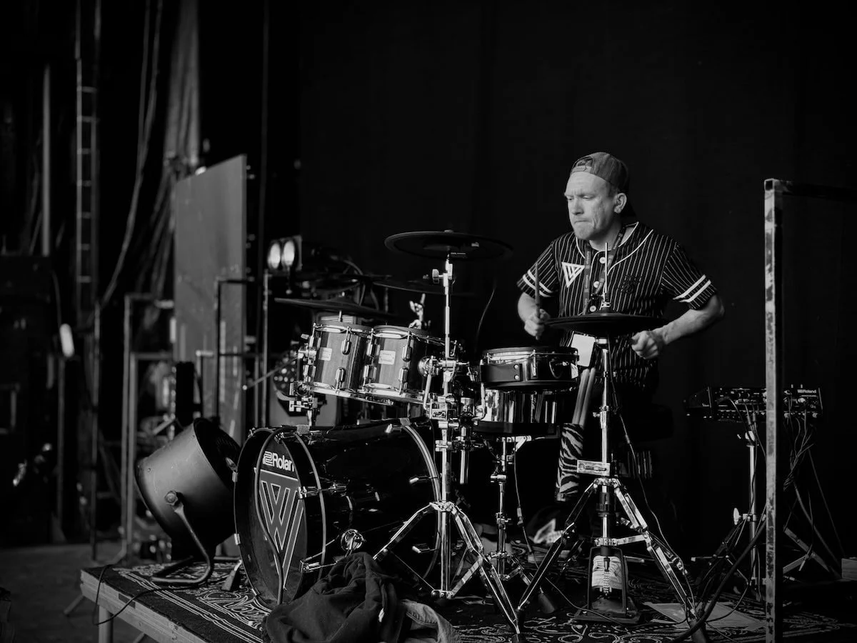 Trummisen från Dunderklubben spelar fokuserat på scen, iförd svart randig tröja och keps bakåtvänd, omgiven av trumset och scenutrustning.