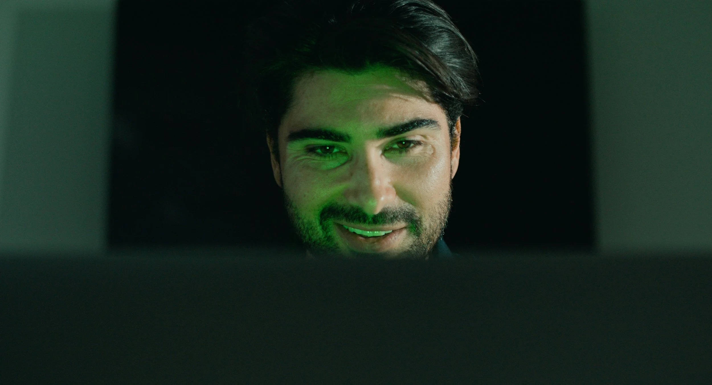 A man with dark hair and a beard looks at a computer screen, with green light reflecting on his face, in a dimly lit room.