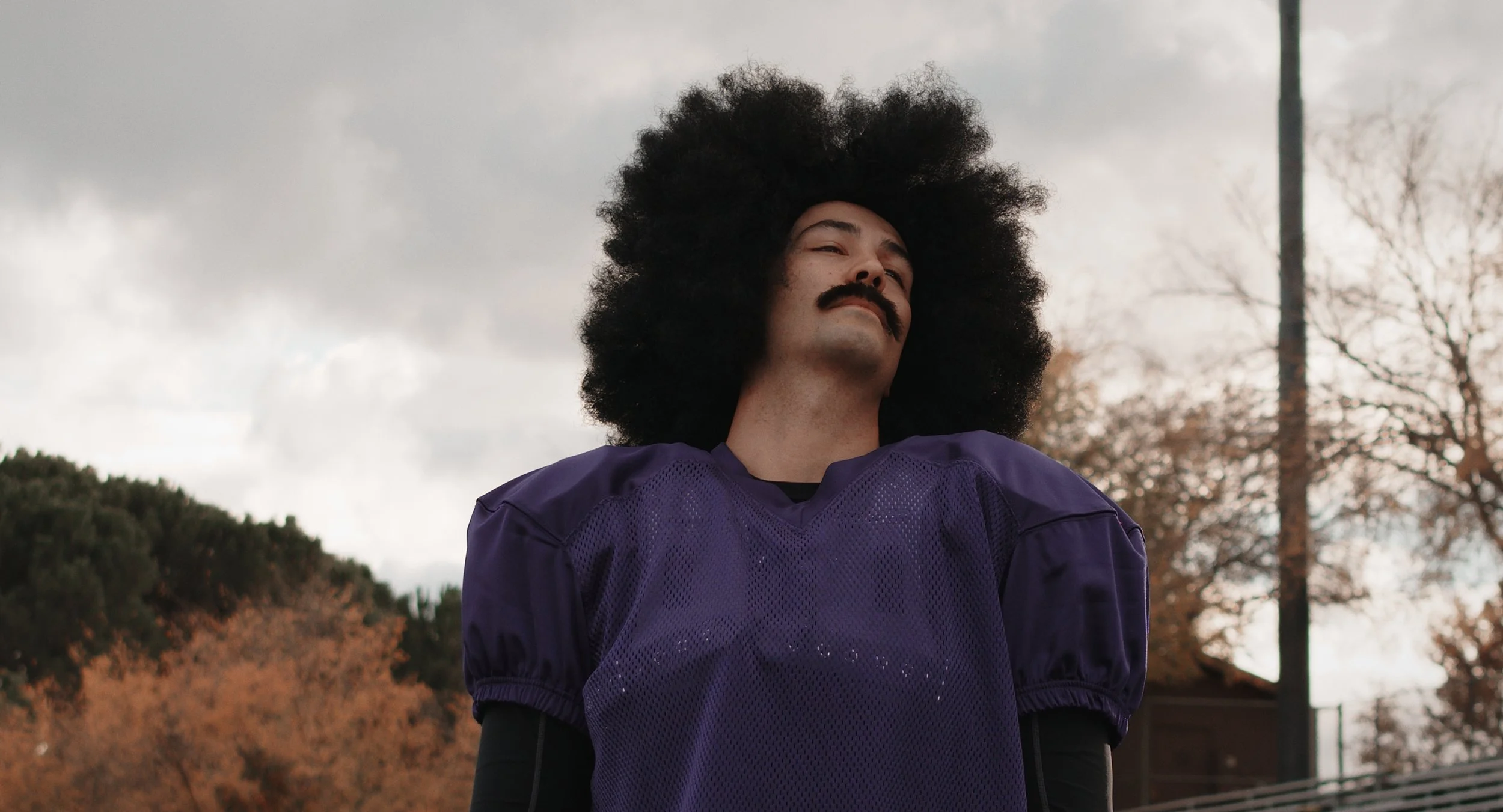 A man with a large afro hairstyle and mustache standing outdoors on a cloudy day, wearing a purple sports jersey.