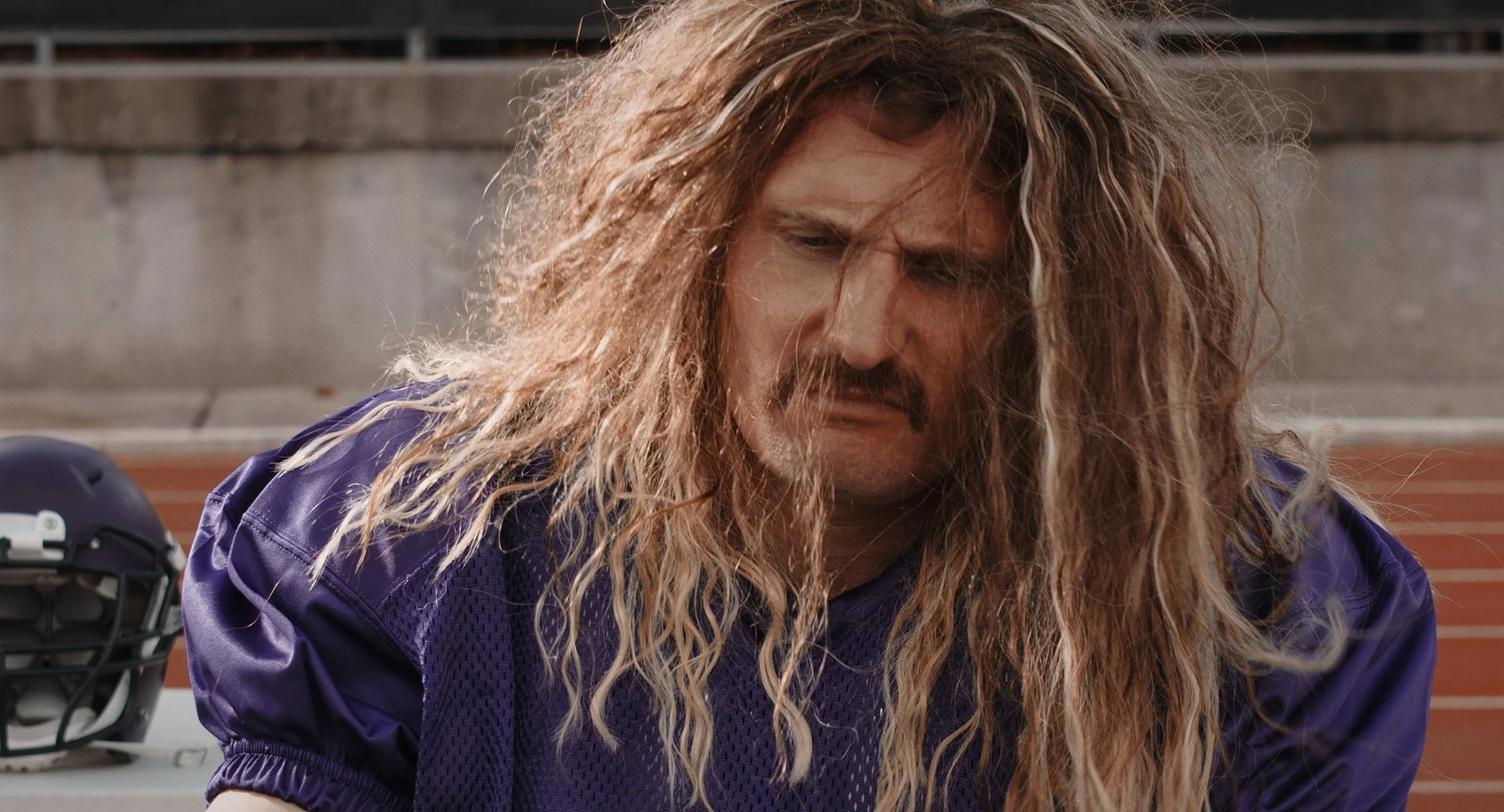A man with long curly hair and a mustache, wearing a purple sports jersey, looks down with a disappointed or anguished expression on a sports field, with a football helmet nearby.