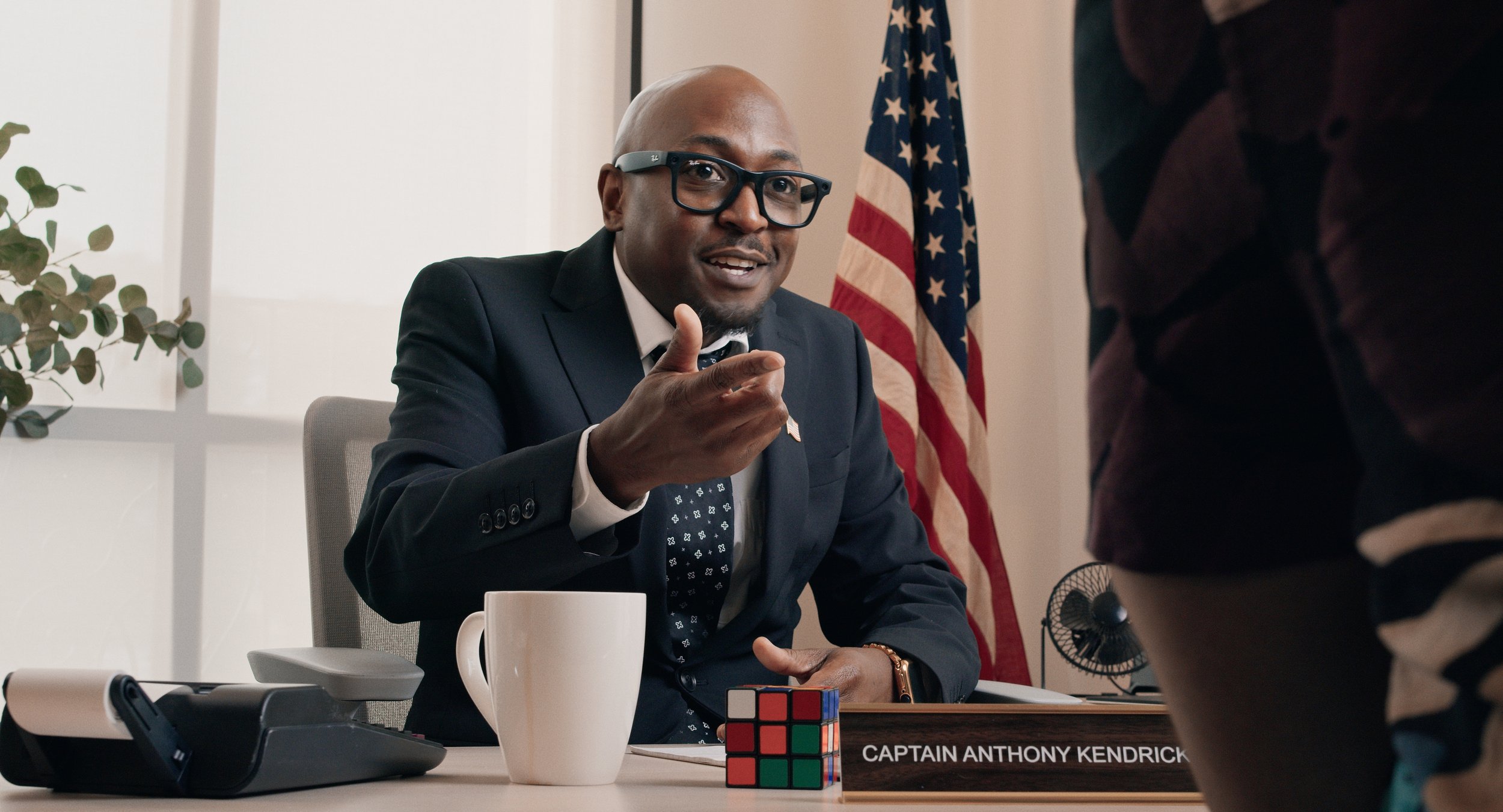 A man in a business suit sitting at a desk in an office, pointing and talking to someone outside the frame. He has a bald head, glasses, and a nameplate on the desk that reads 'Captain Anthony Kendrick'. There is an American flag behind him, a coffee
