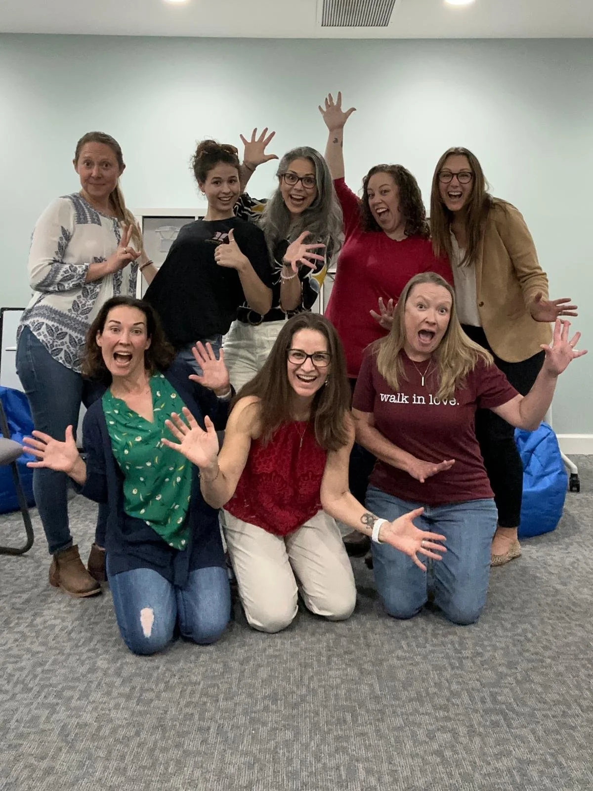 Group of eight women posing playfully indoors, some kneeling and some standing, all with funny expressions and hand gestures.