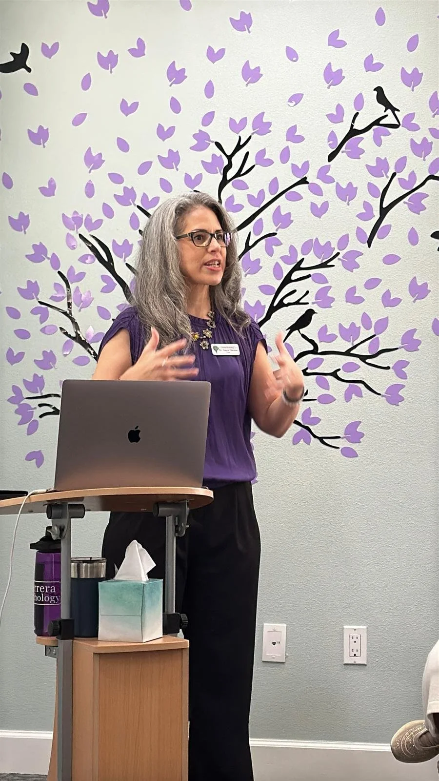 A woman with gray hair and glasses is speaking in front of a podium with an open laptop. She is gesturing with her hands. Behind her, there is a wall mural of a tree with purple leaves and black branches, with bird silhouettes. On the podium, there are a tissue box and a couple of drink containers.