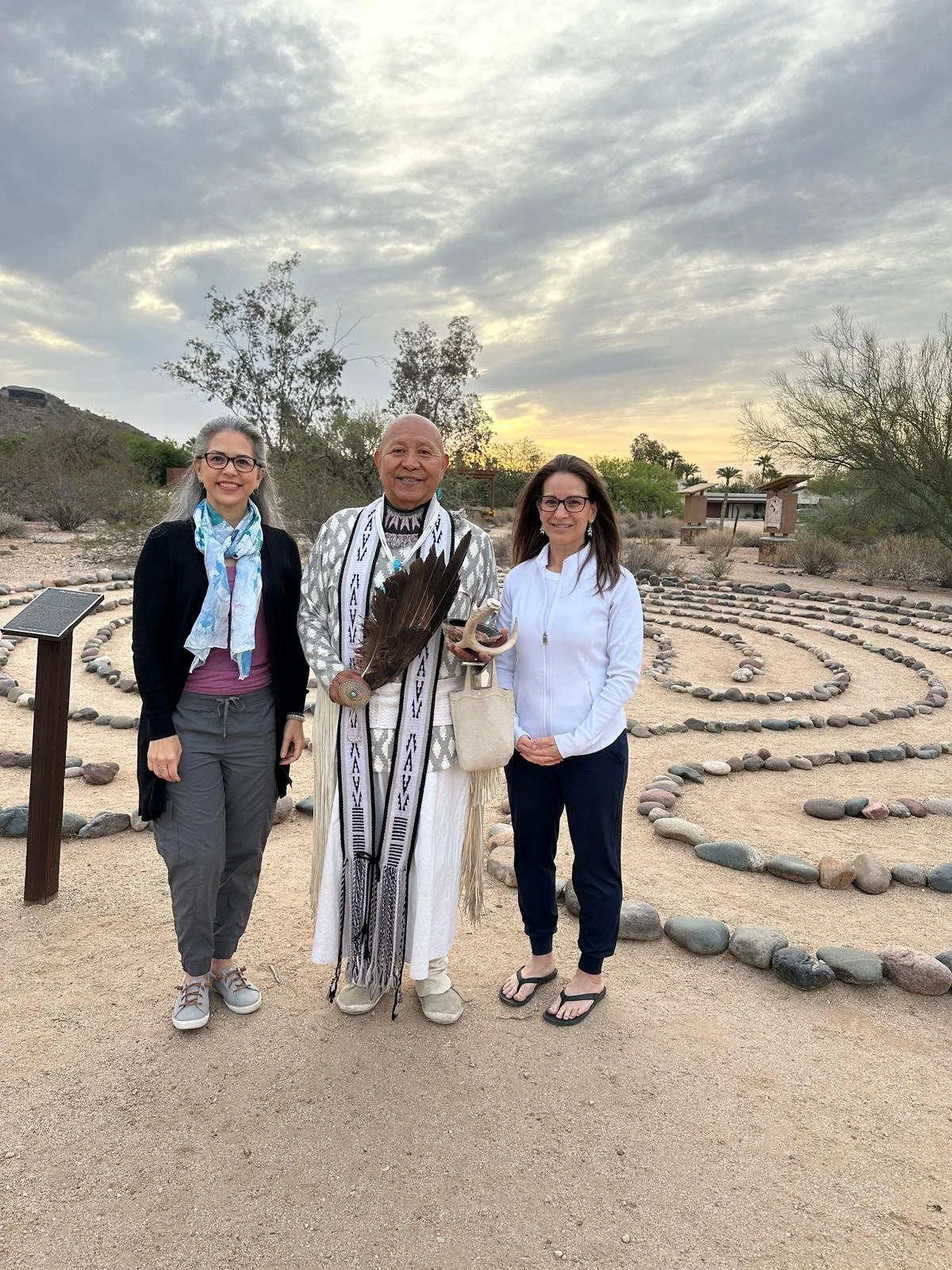 Three people standing together outdoors in a desert landscape with a cloudy sky. One person is wearing traditional attire and holding feathers, while the other two are dressed in casual clothing. The background features a rock path winding through the sand and scattered vegetation, with mountains in the distance.