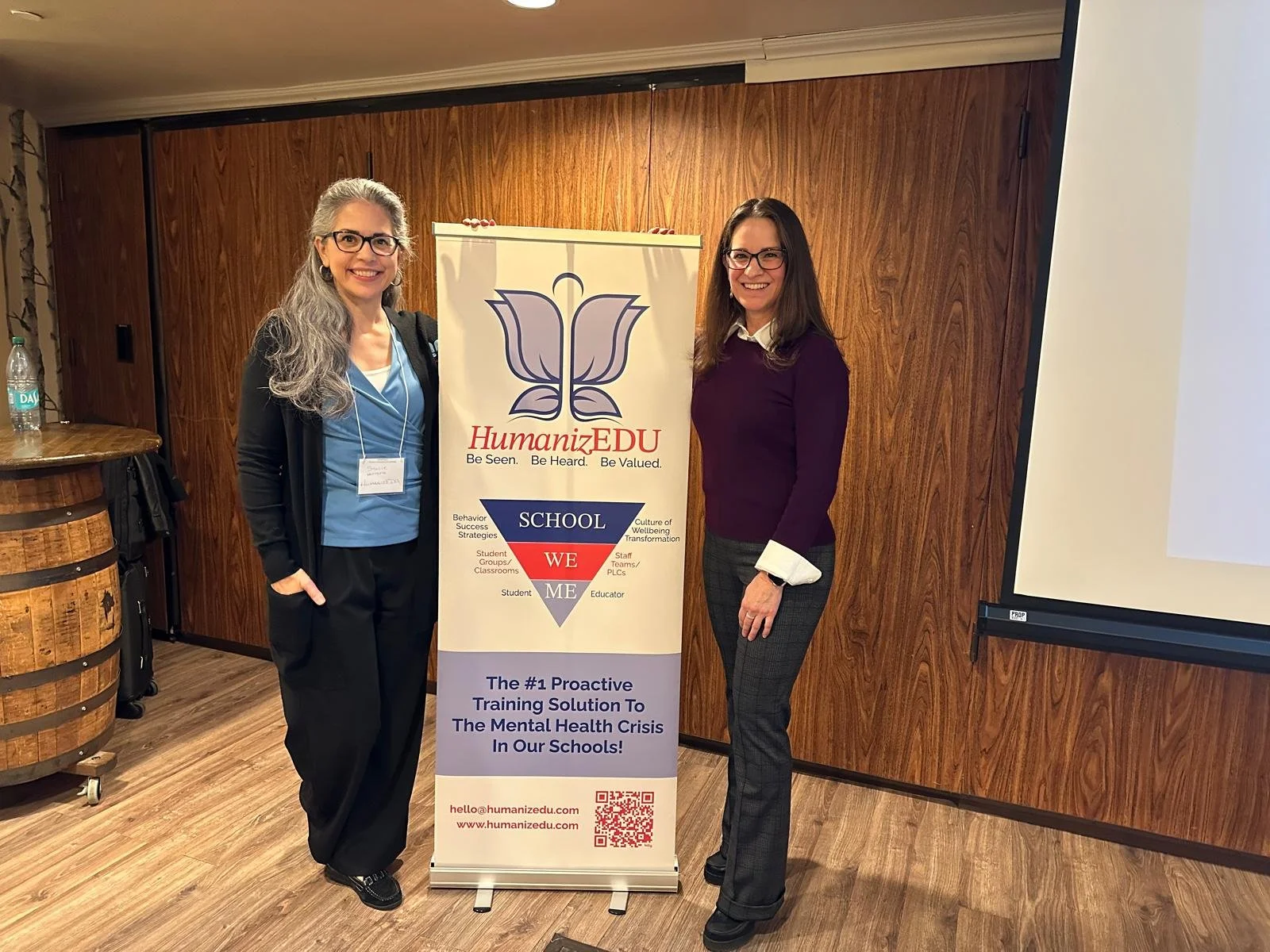 Two women standing beside a HumanizEDU banner in an indoor setting with a wooden backdrop and projector screen. The banner highlights mental health training solutions for schools.
