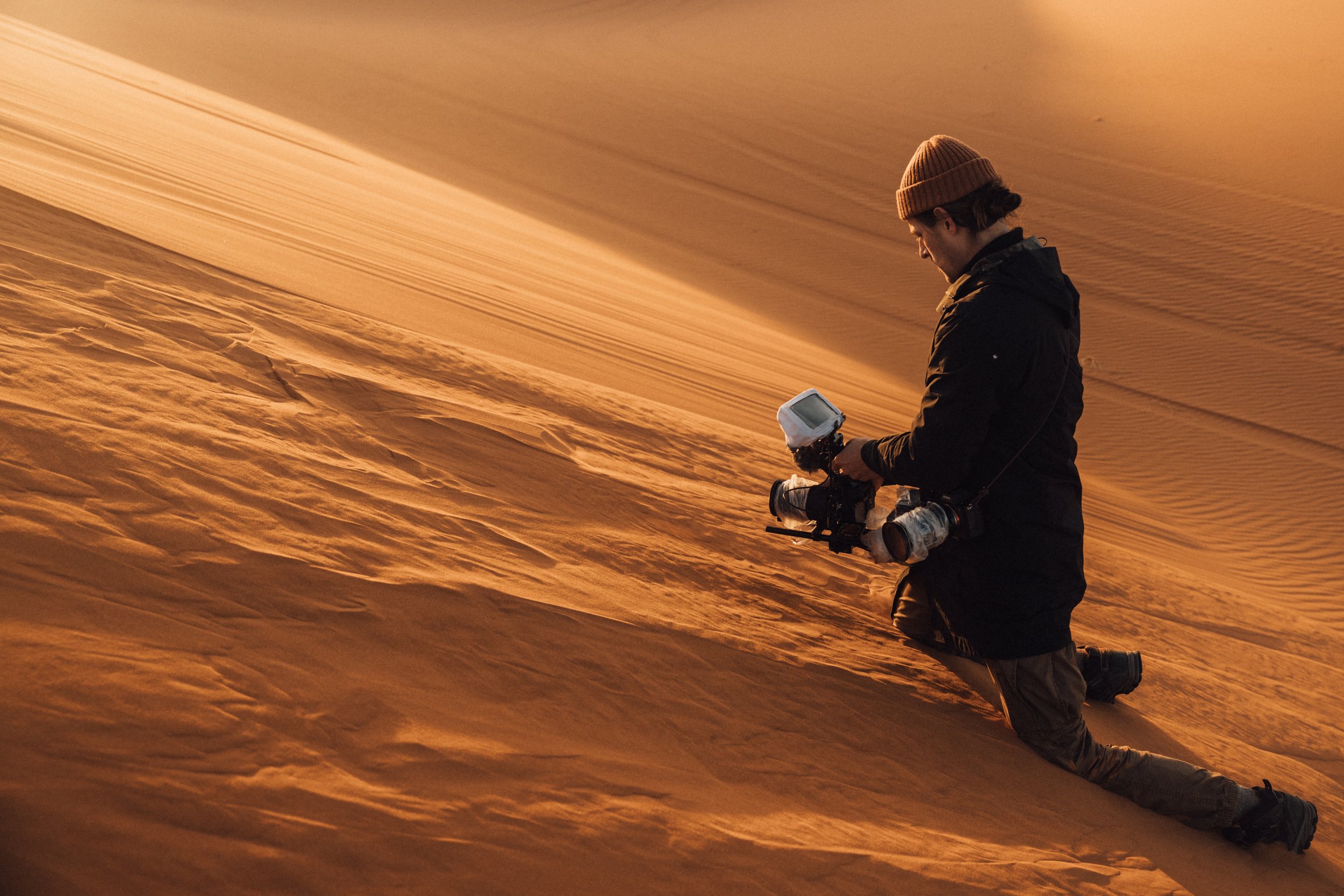 Person kneeling on desert sand dunes, taking photographs with a camera, at sunset, wearing a beanie and dark jacket.