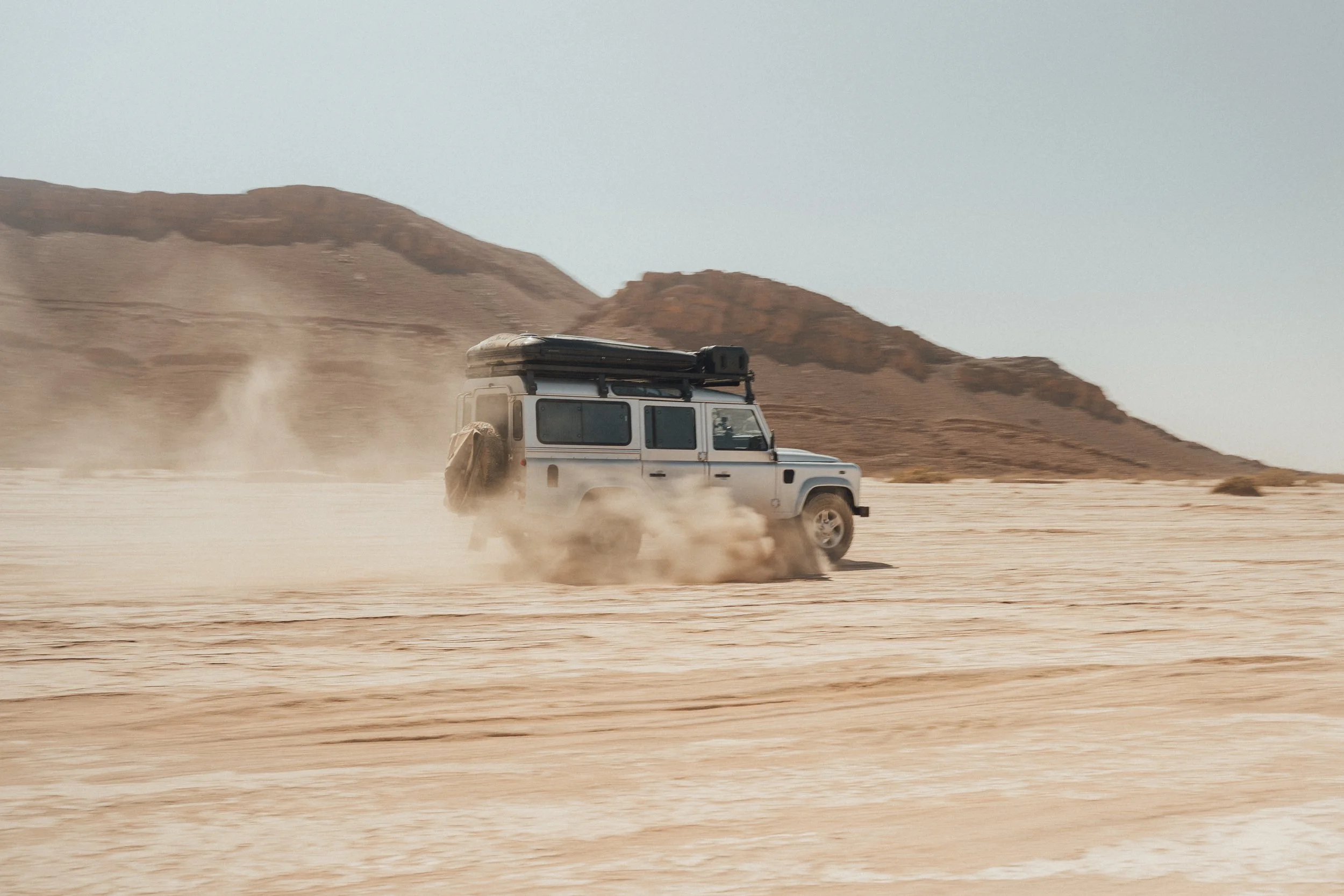 A white off-road vehicle driving across a desert landscape, kicking up dust with mountains in the background.