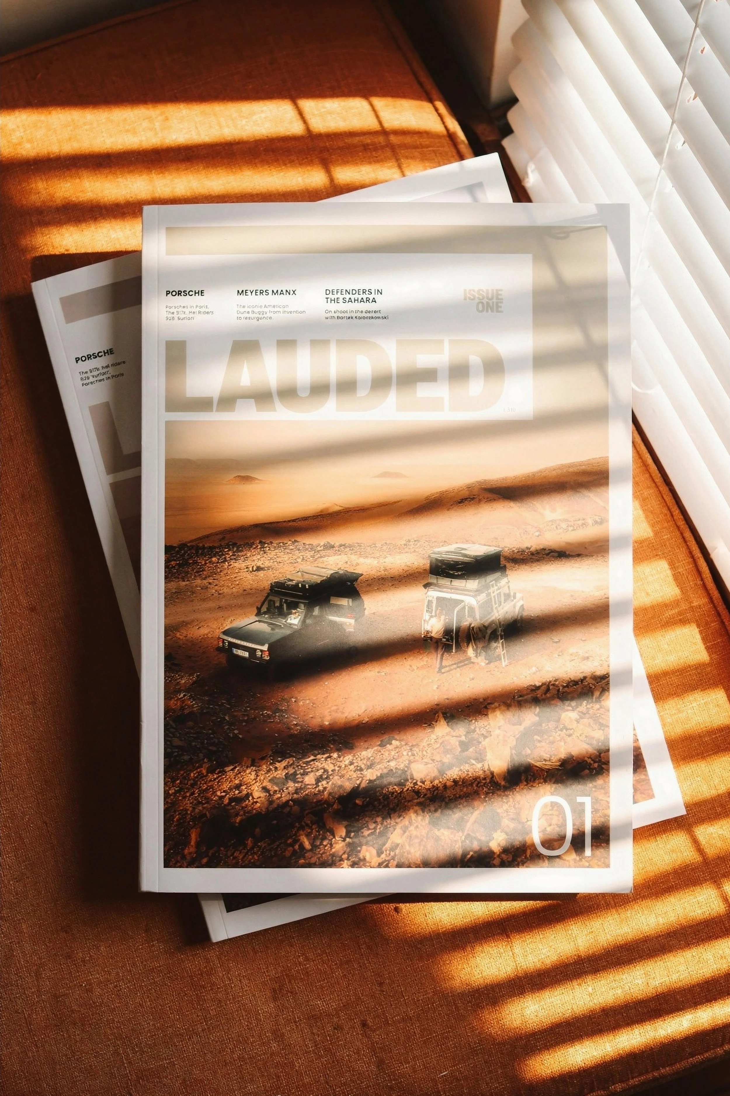 Two magazines or books stacked on a burnt orange cushion with blinds casting shadows on them. The top magazine features an image of a desert landscape with two off-road vehicles and the word 'LAUDED' in large letters. The cover also includes headline
