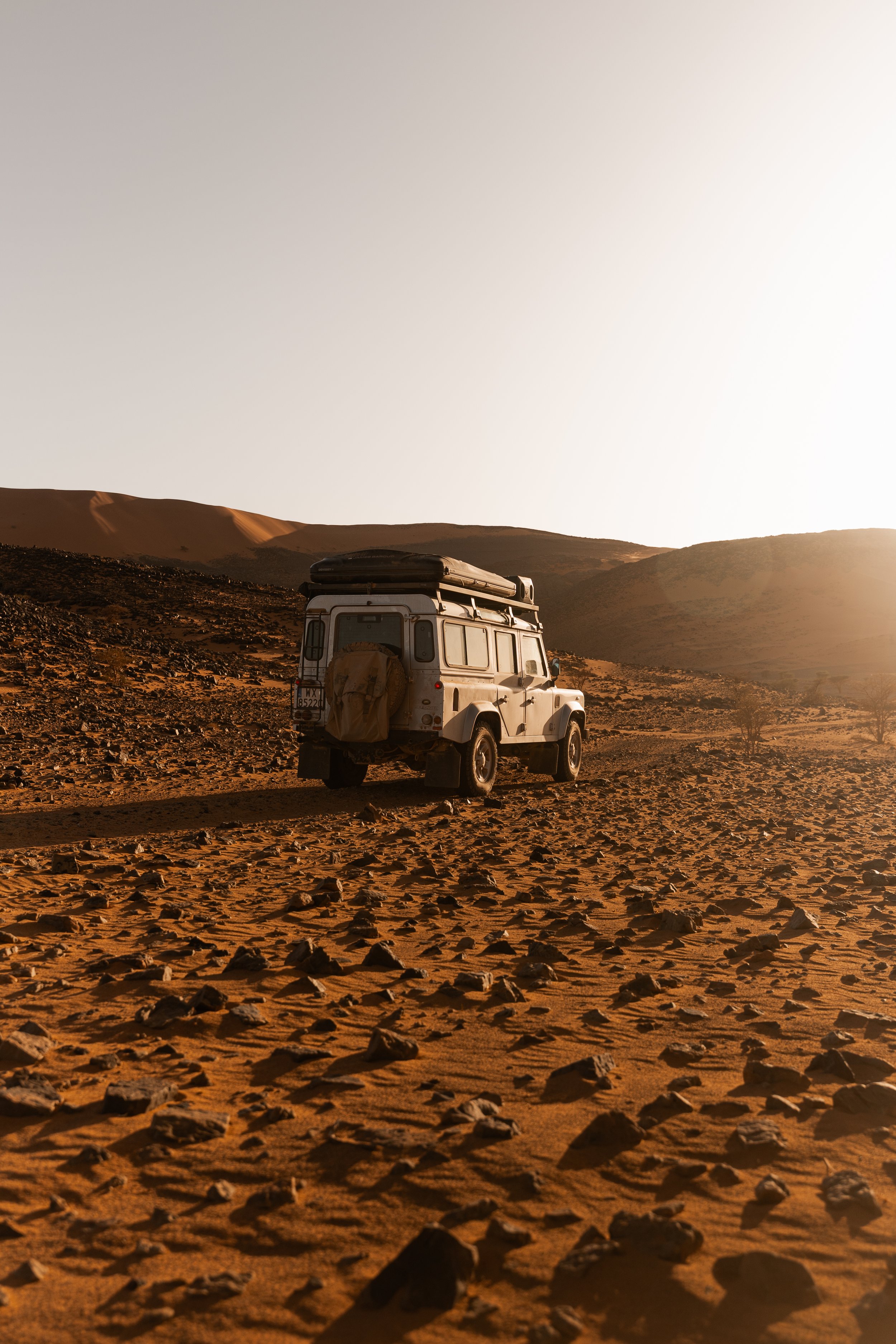 A white off-road vehicle with a roof storage box and a covered spare tire in a desert landscape during sunset, with sand dunes and rocky terrain.