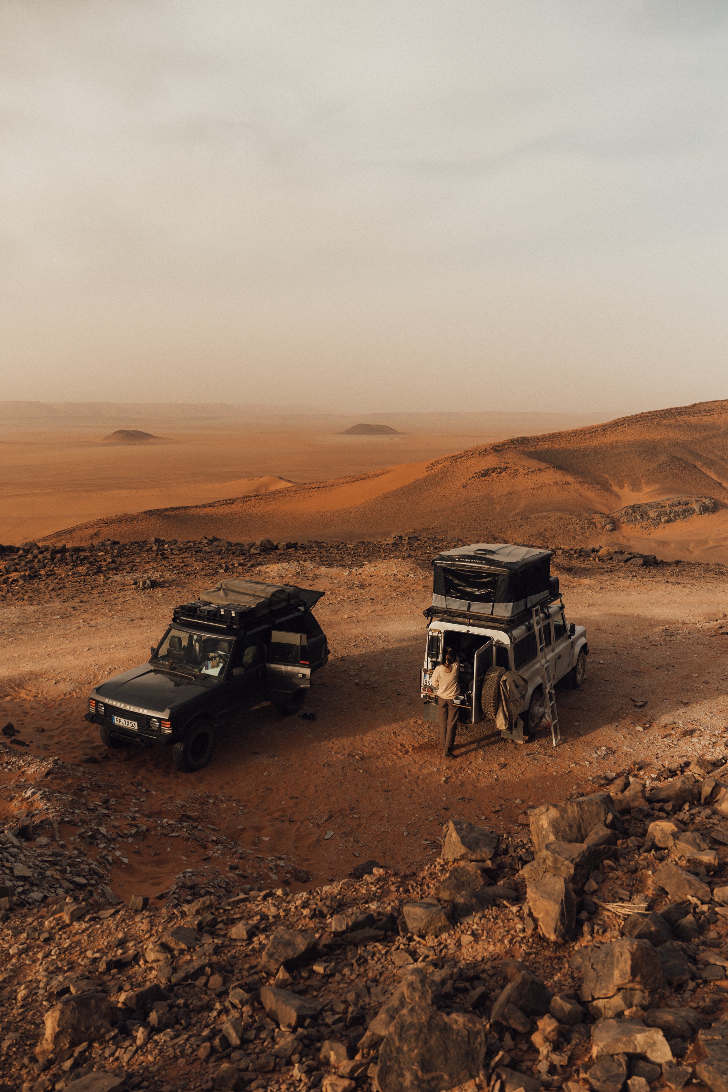 Two off-road vehicles parked on a rocky desert landscape with a person standing beside one of the vehicles, overlooking a vast, barren desert terrain.